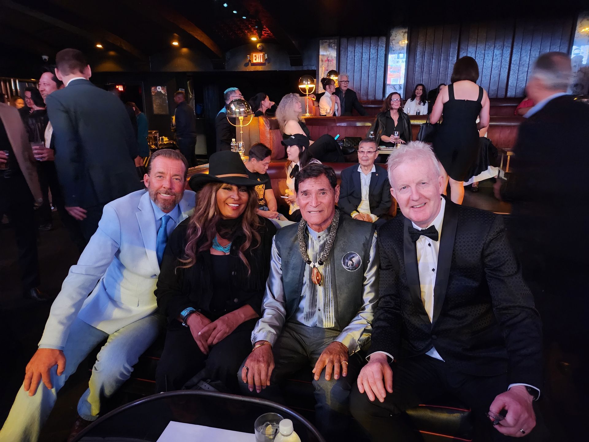 Four people seated together at a formal event, smiling at the camera. They are in a dimly lit lounge with other attendees.