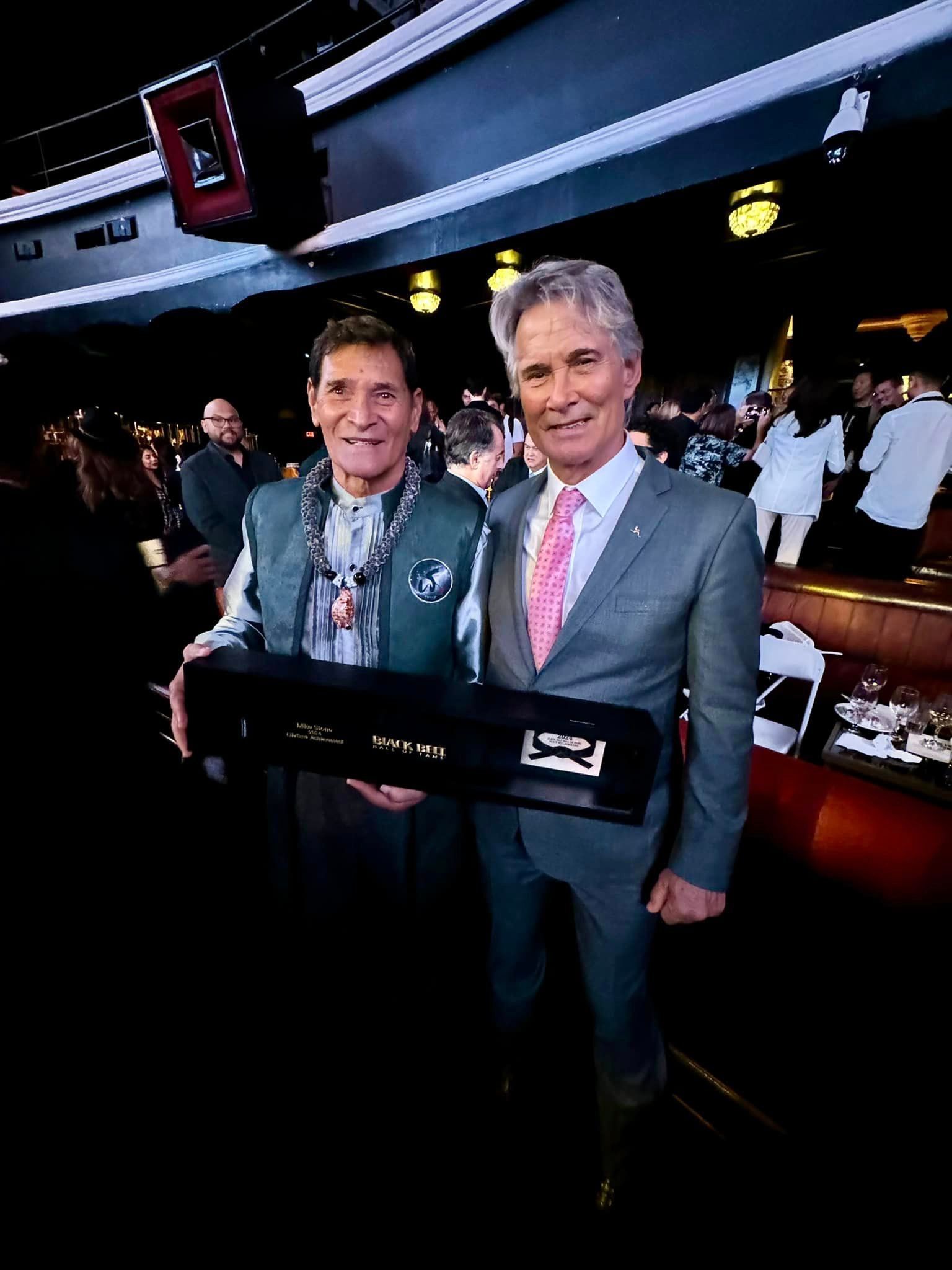 Two men pose with a case in a venue. The man on the left wears a vest and the other, a suit.