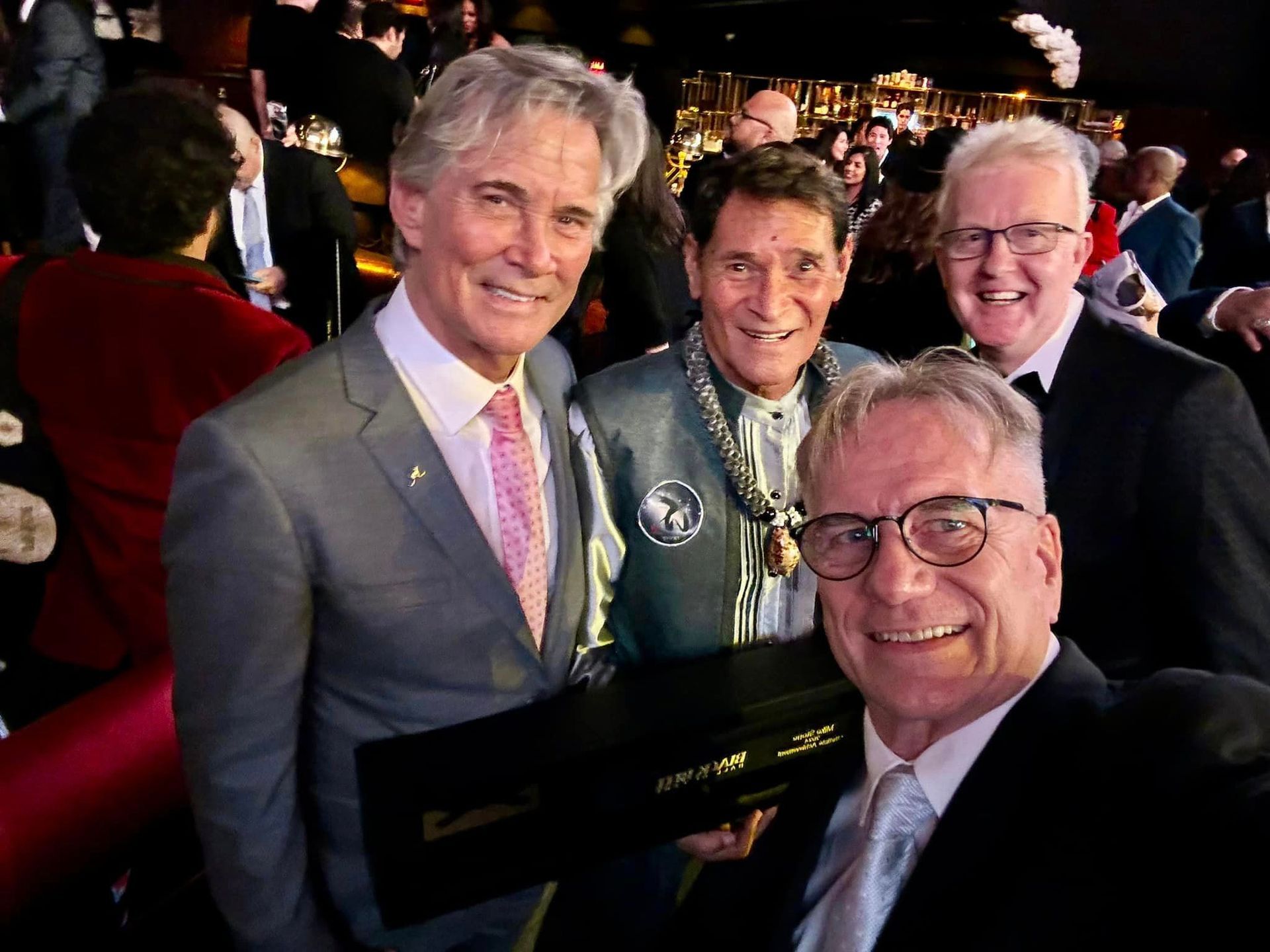Four men smile for a selfie at an event. One man holds a black box, another wears a medallion.