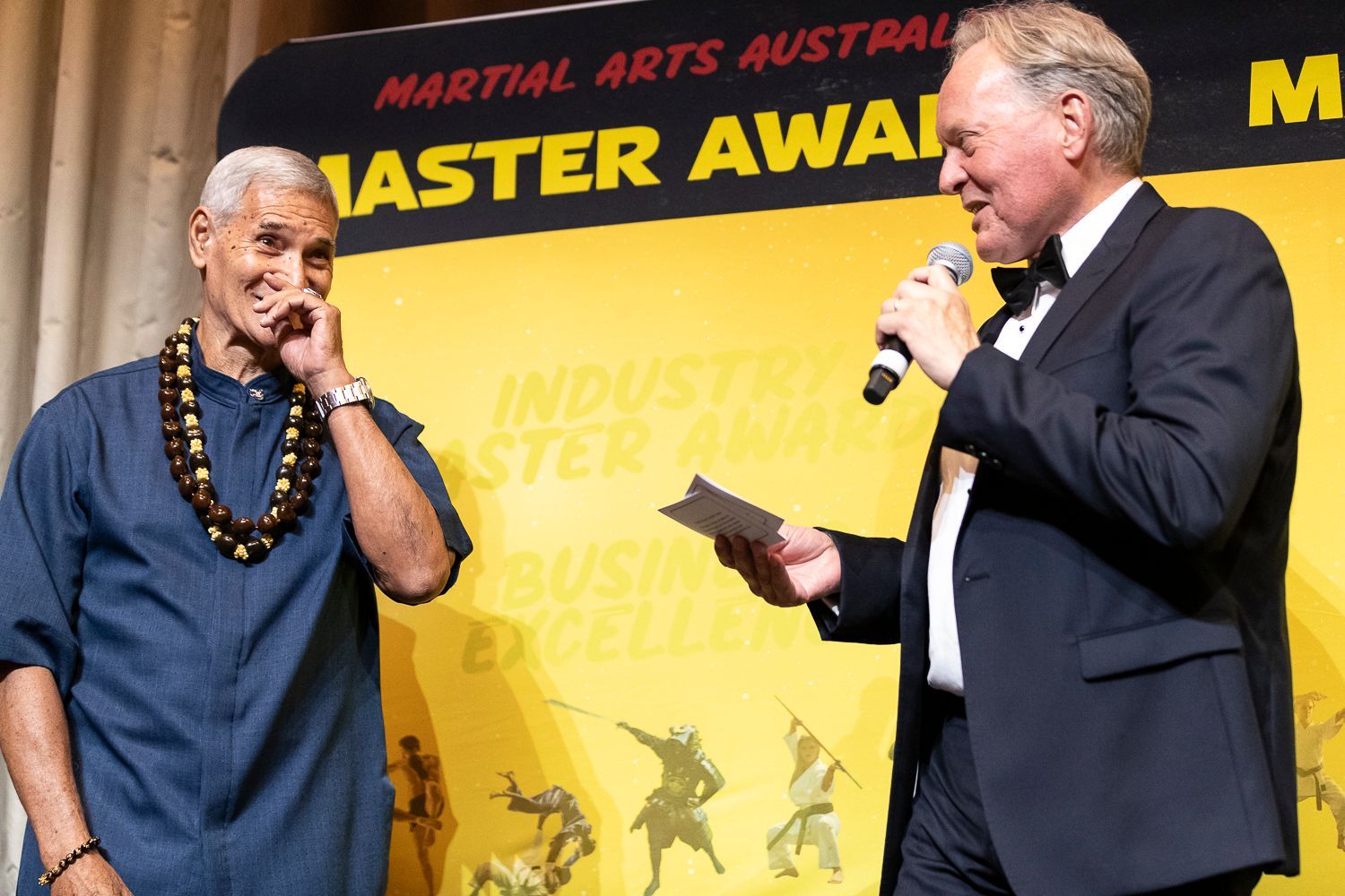 Two men at an awards ceremony: one in a black suit with a microphone.