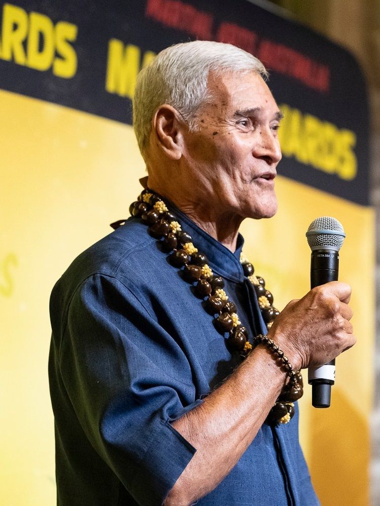 Man with gray hair, wearing lei and holding a microphone, speaking.