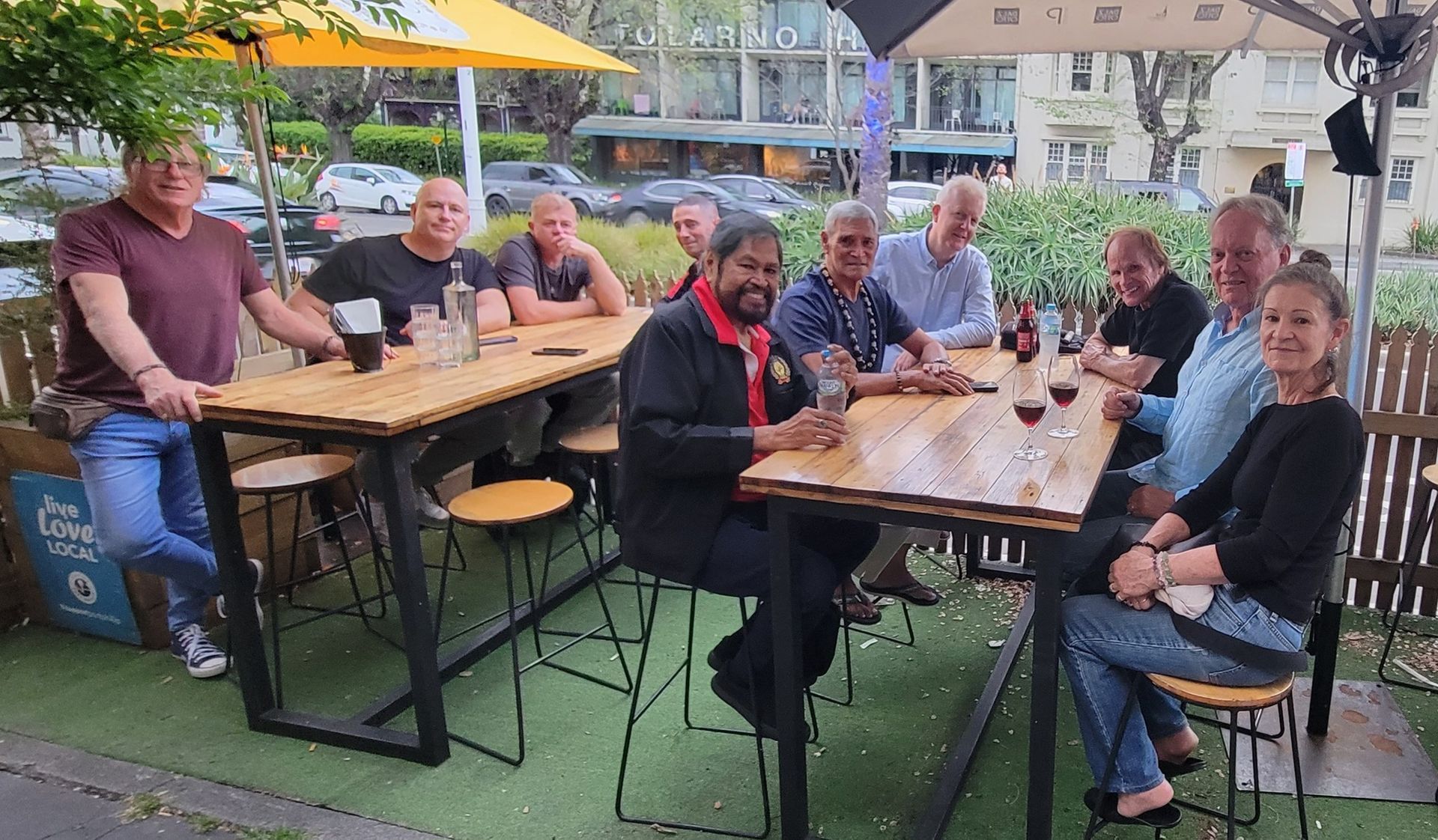 A group of ten people seated around two wooden tables outside a cafe.