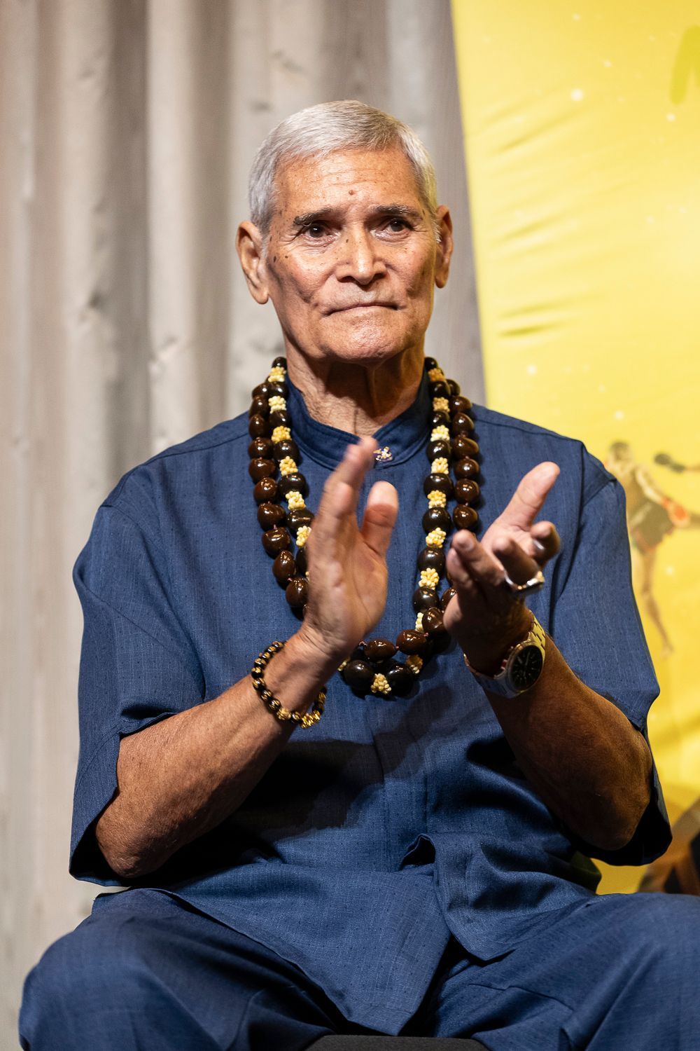 An older man with gray hair, wearing a lei and blue shirt, clapping his hands.