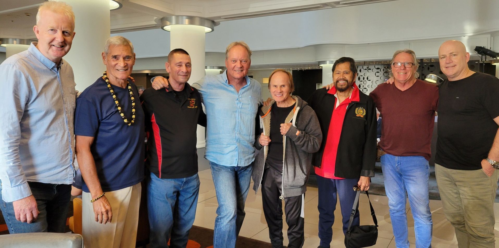 Eight men of diverse ethnicities pose closely together in an indoor setting, possibly a lobby.