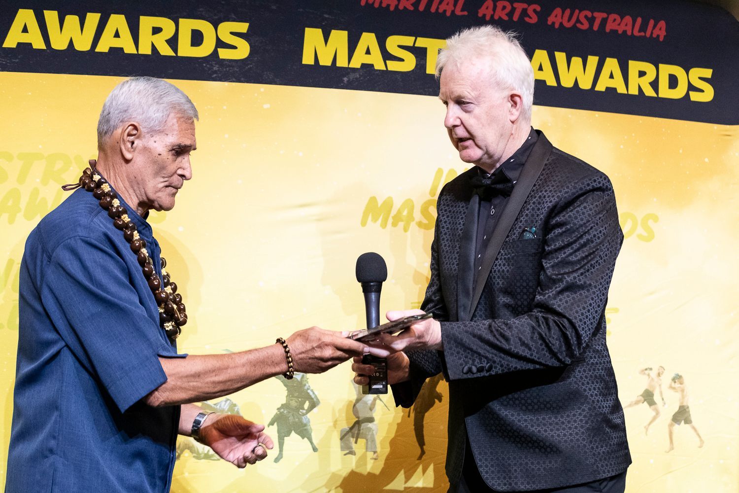 Two men on stage at an awards ceremony, one presenting the other with a plaque; backdrop says 