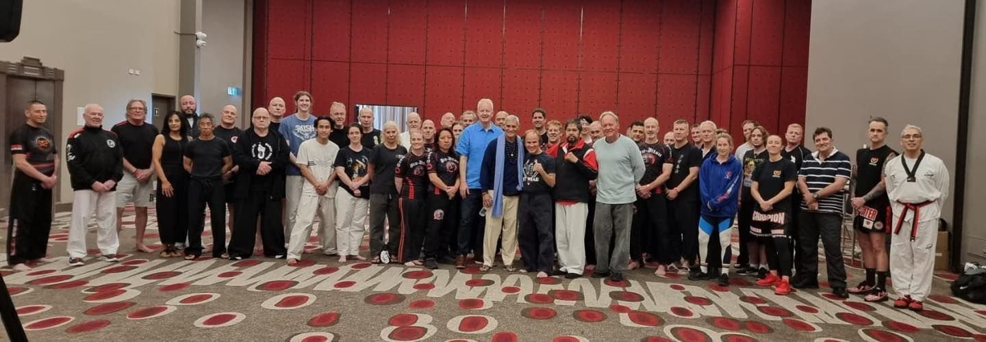 A large group of people in martial arts uniforms pose in a conference room.  Red carpet and backdrop.