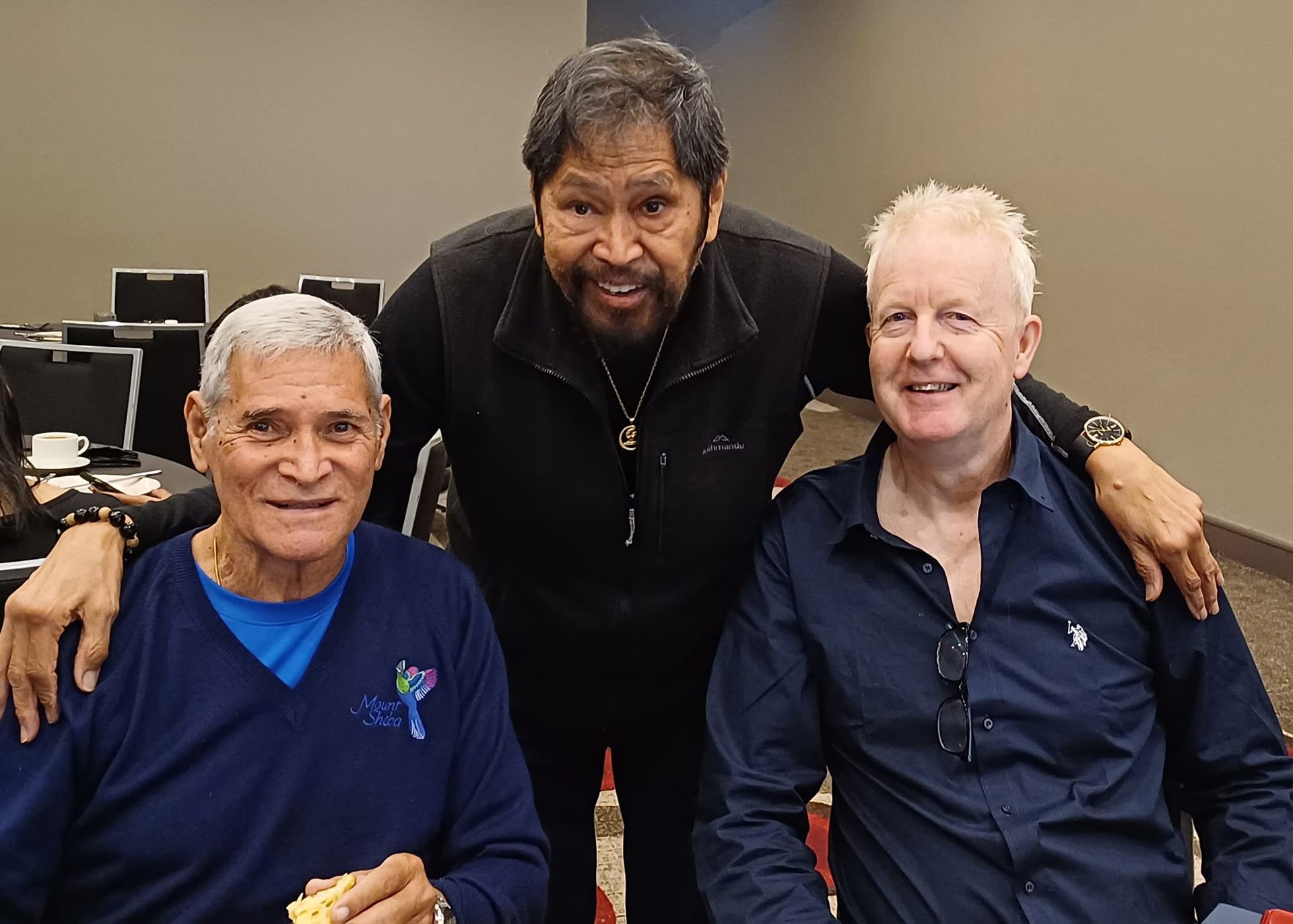 Three men posing for a photo. One man has arms around the other two. All have smiles. The setting appears to be indoors.
