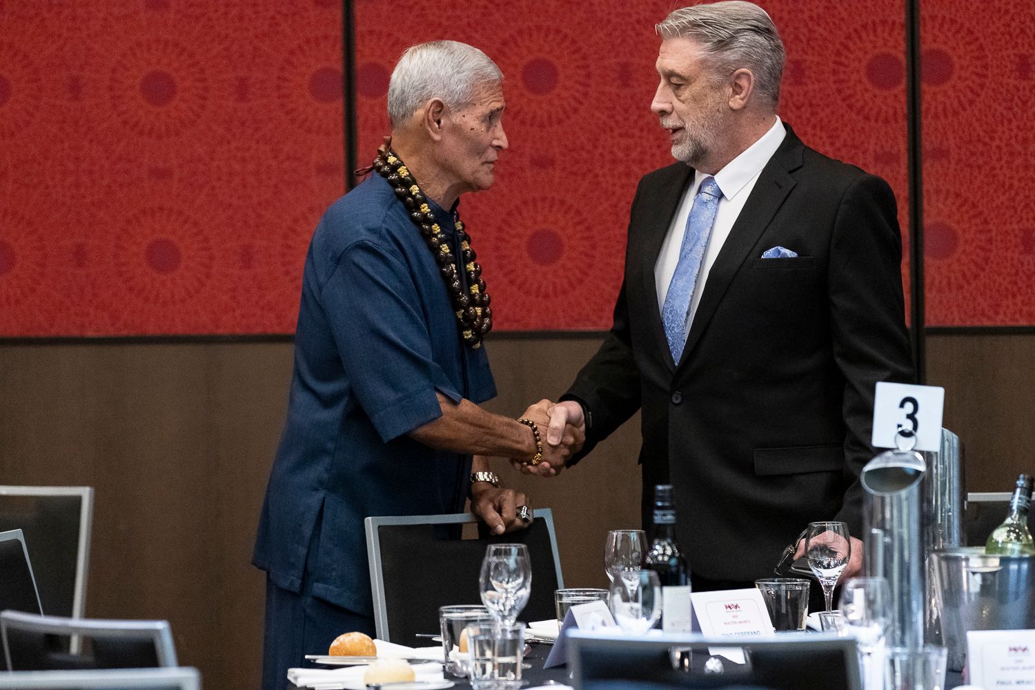 Two men shaking hands at a formal event, one in a blue suit and tie.