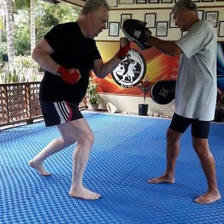 Two older men in a gym: one throwing a punch at a mitt held by the other. Both barefoot, on a blue mat.