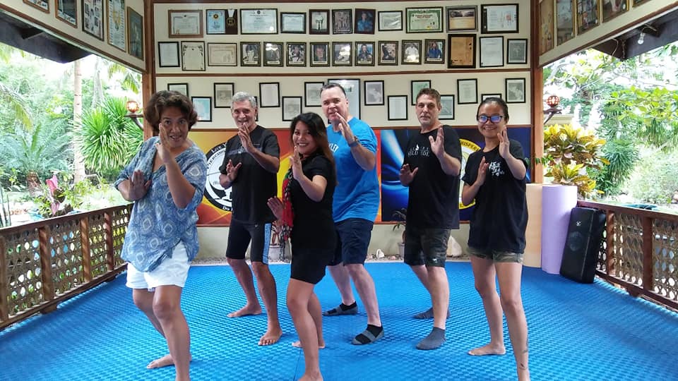 Six people in a pavilion, practicing martial arts. They stand in a line, striking poses, with framed awards on the walls.