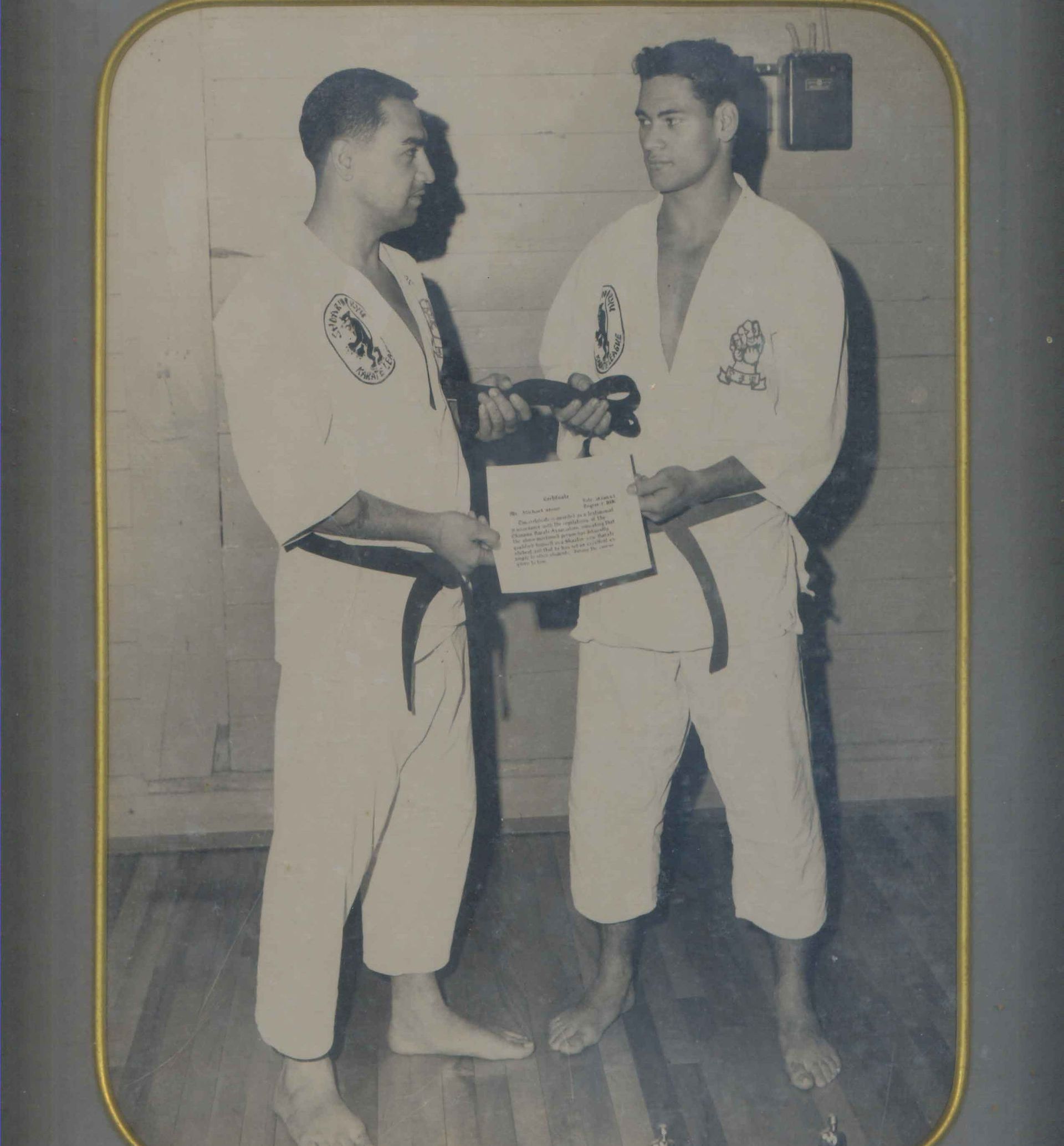 Two men in karate uniforms, one handing the other a document. Black and white photo, indoor setting.