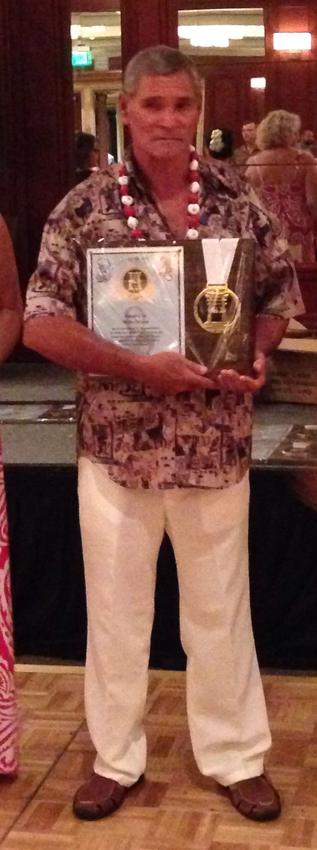 Man in floral shirt and white pants holding awards, wearing a lei. Brown shoes, inside setting.