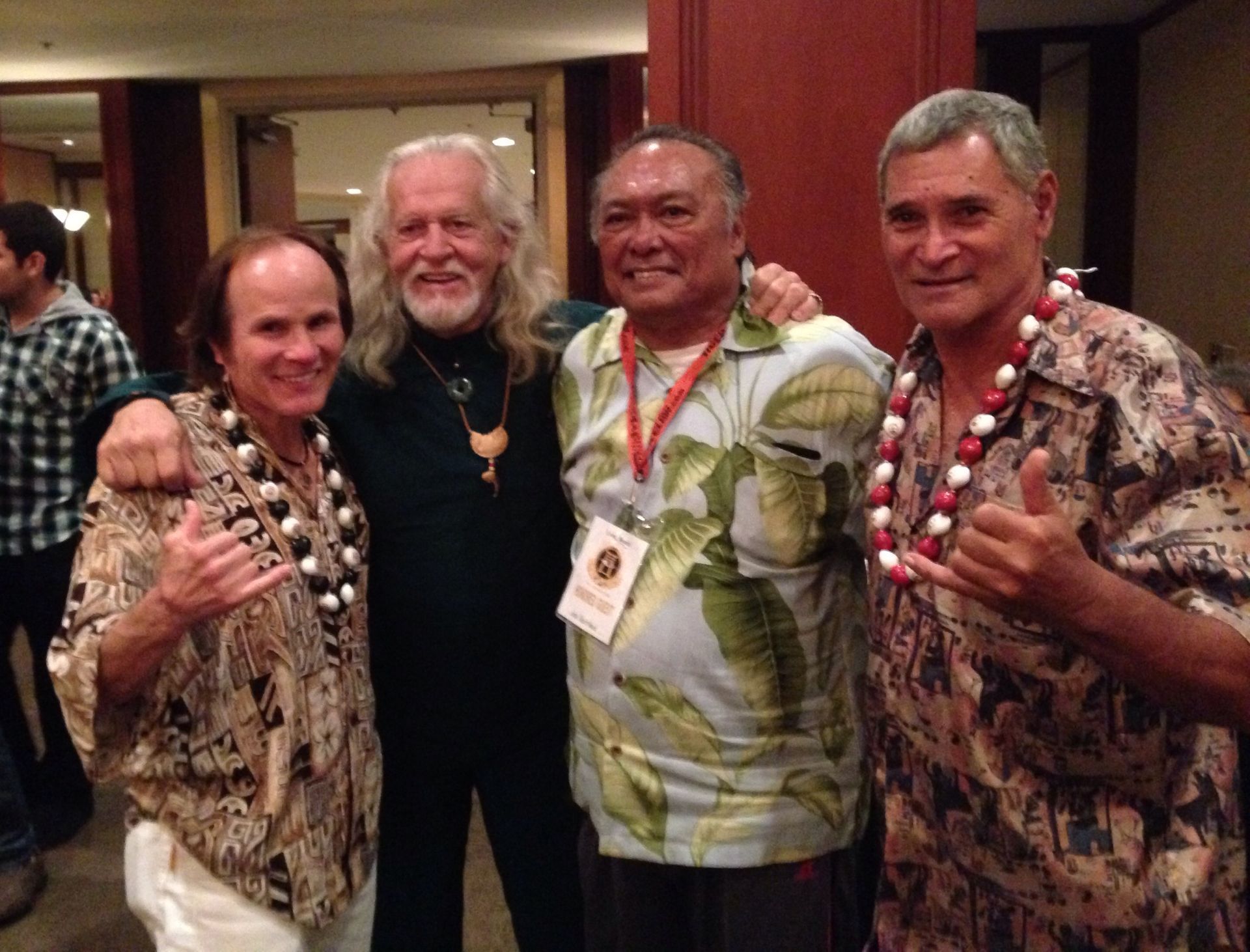 Four men wearing leis and aloha shirts pose for a photo indoors. They are smiling and giving shaka signs.