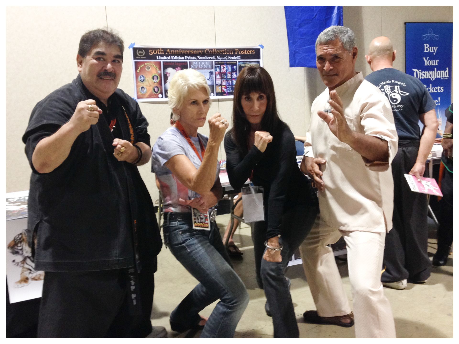 Four people posing in martial arts stances. They are indoors, with posters and other people in the background.