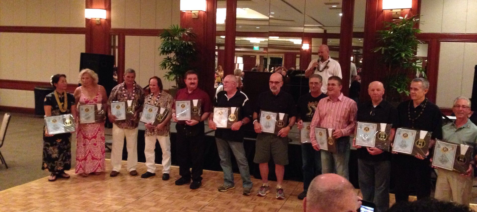 A group of people holding framed awards at an event. The room has wooden accents and some plants. 