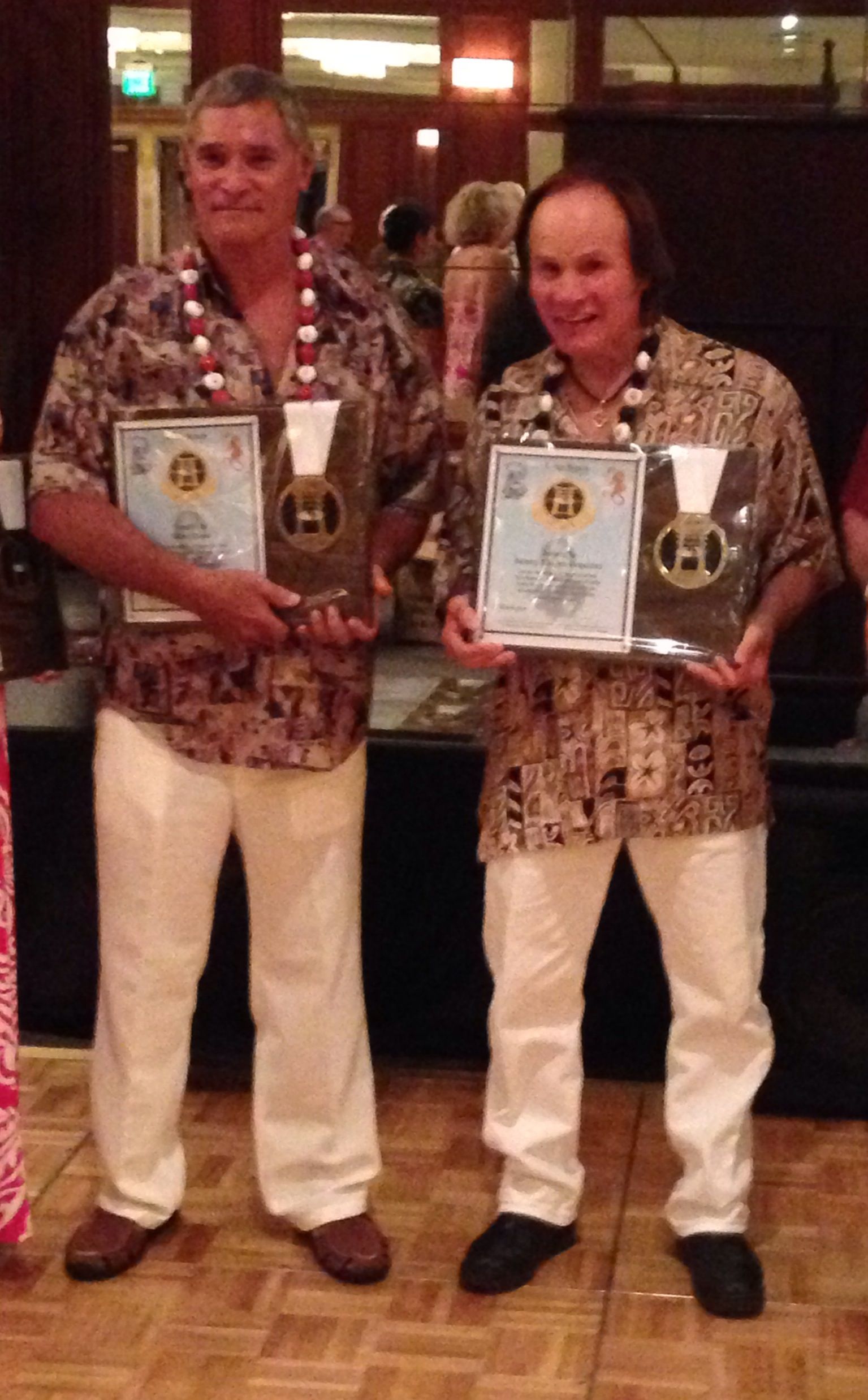 Two men in Hawaiian shirts and white pants holding awards, smiling, indoors.