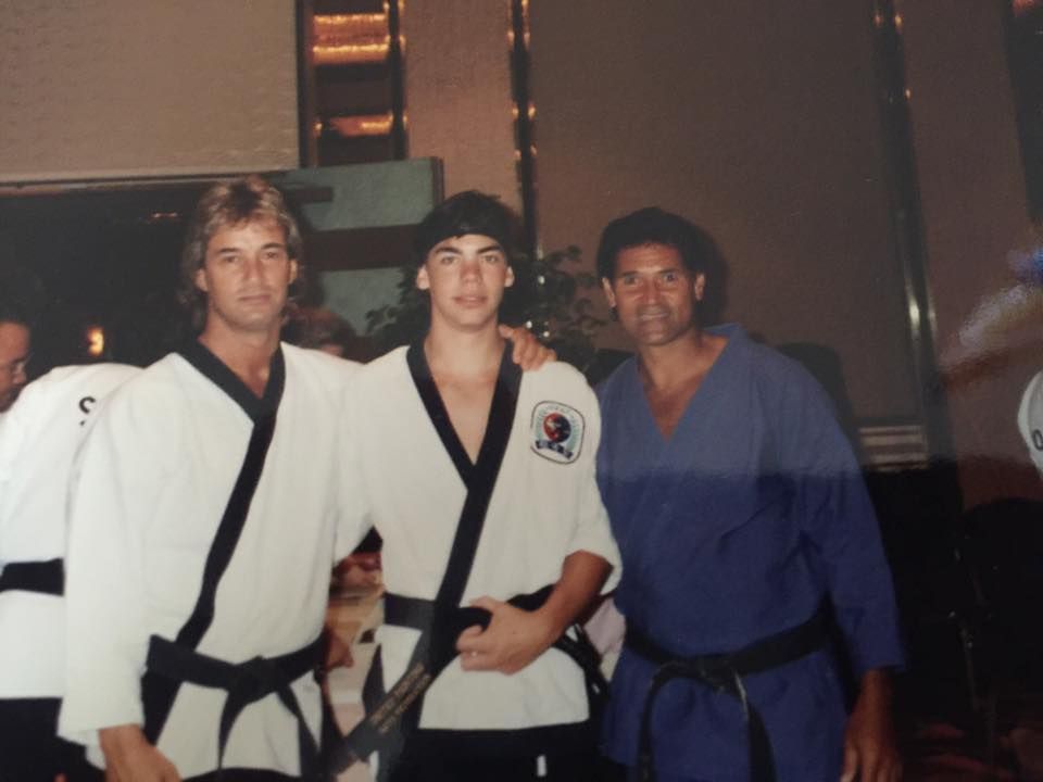 Three men in martial arts uniforms pose indoors; two in white with black belts, the third in blue. They are smiling.