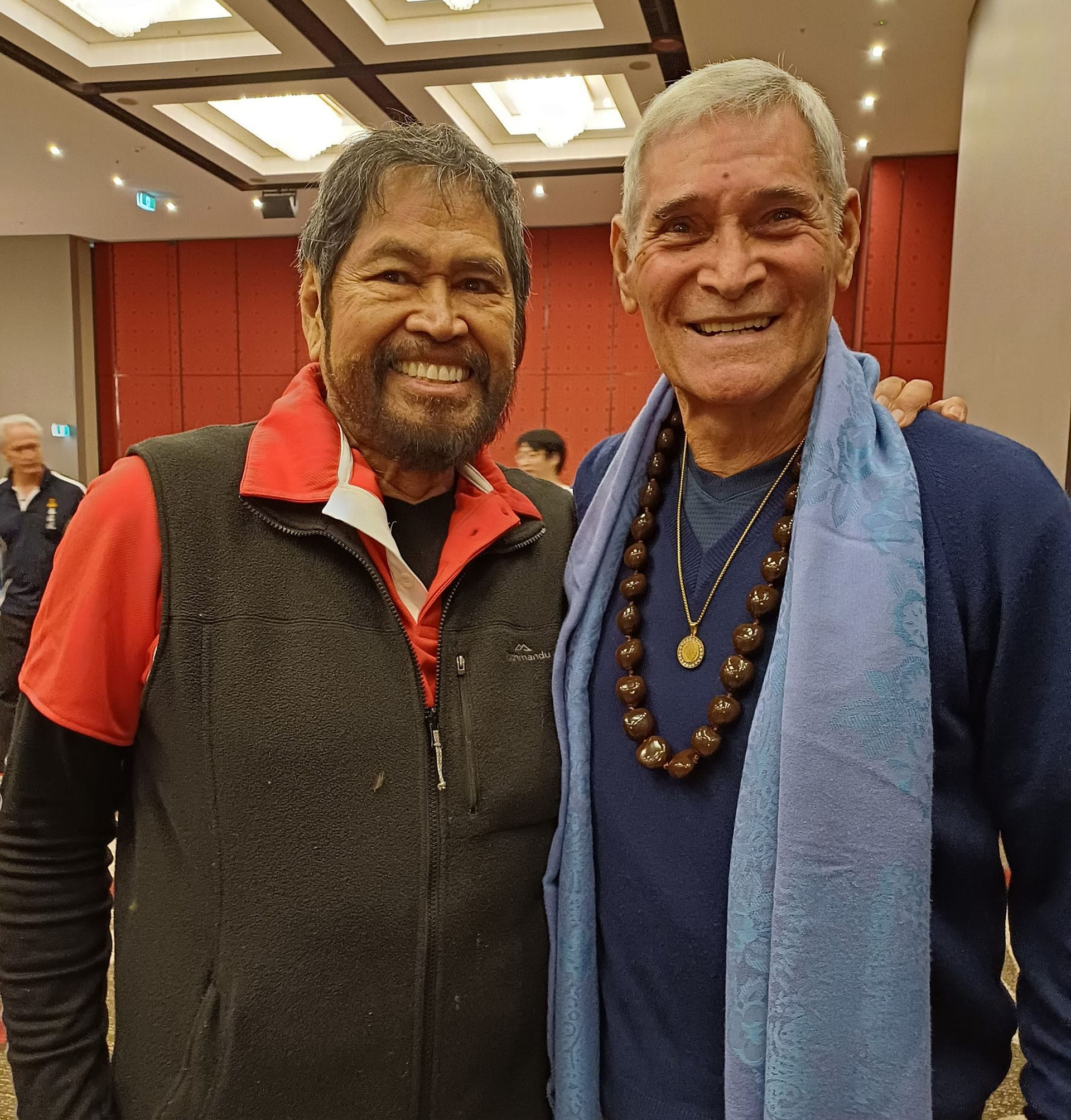 Two men smiling at the camera. One wears a red shirt and vest. The other wears a blue sweater, scarf, and a beaded necklace.
