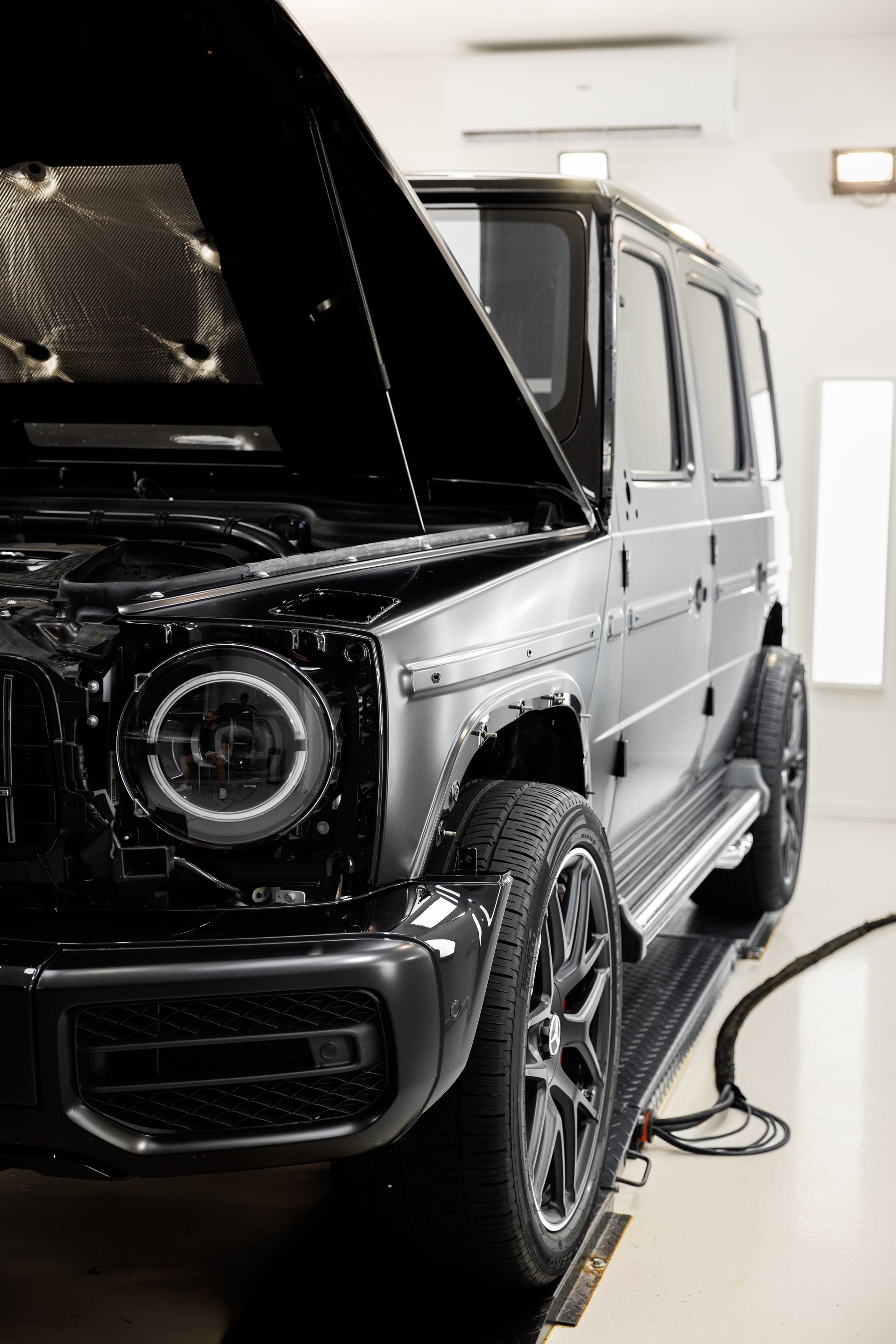 A black Mercedes-Benz G-Wagon with its hood open, parked in a well-lit, professional automotive workshop.