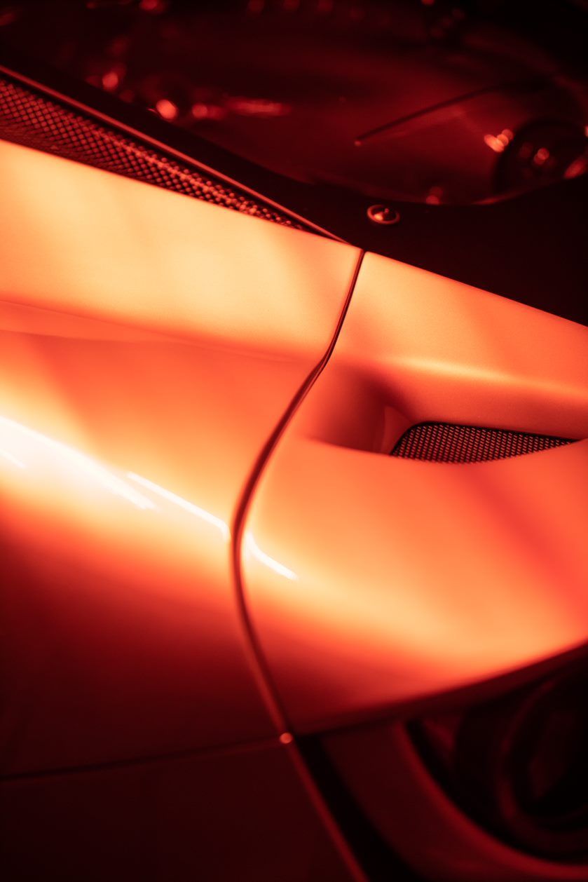 Close-up of a red-lit car's body, showing a curving side panel, a small air vent, and part of the windshield.