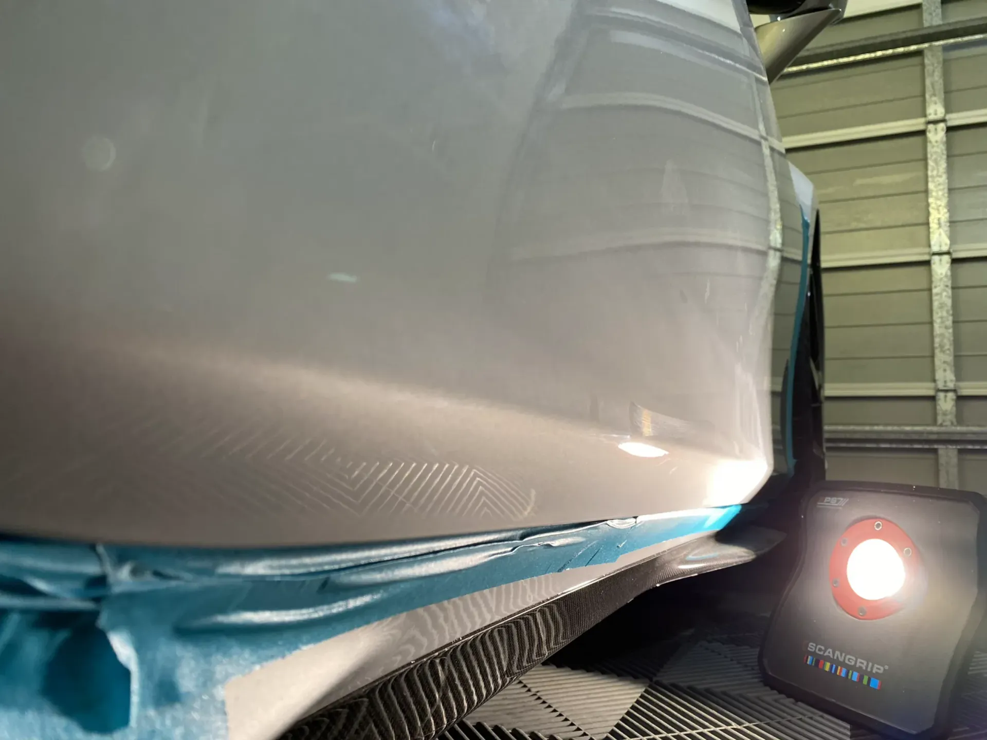 White car bumper being painted, blue tape, workshop setting, and a red spotlight.