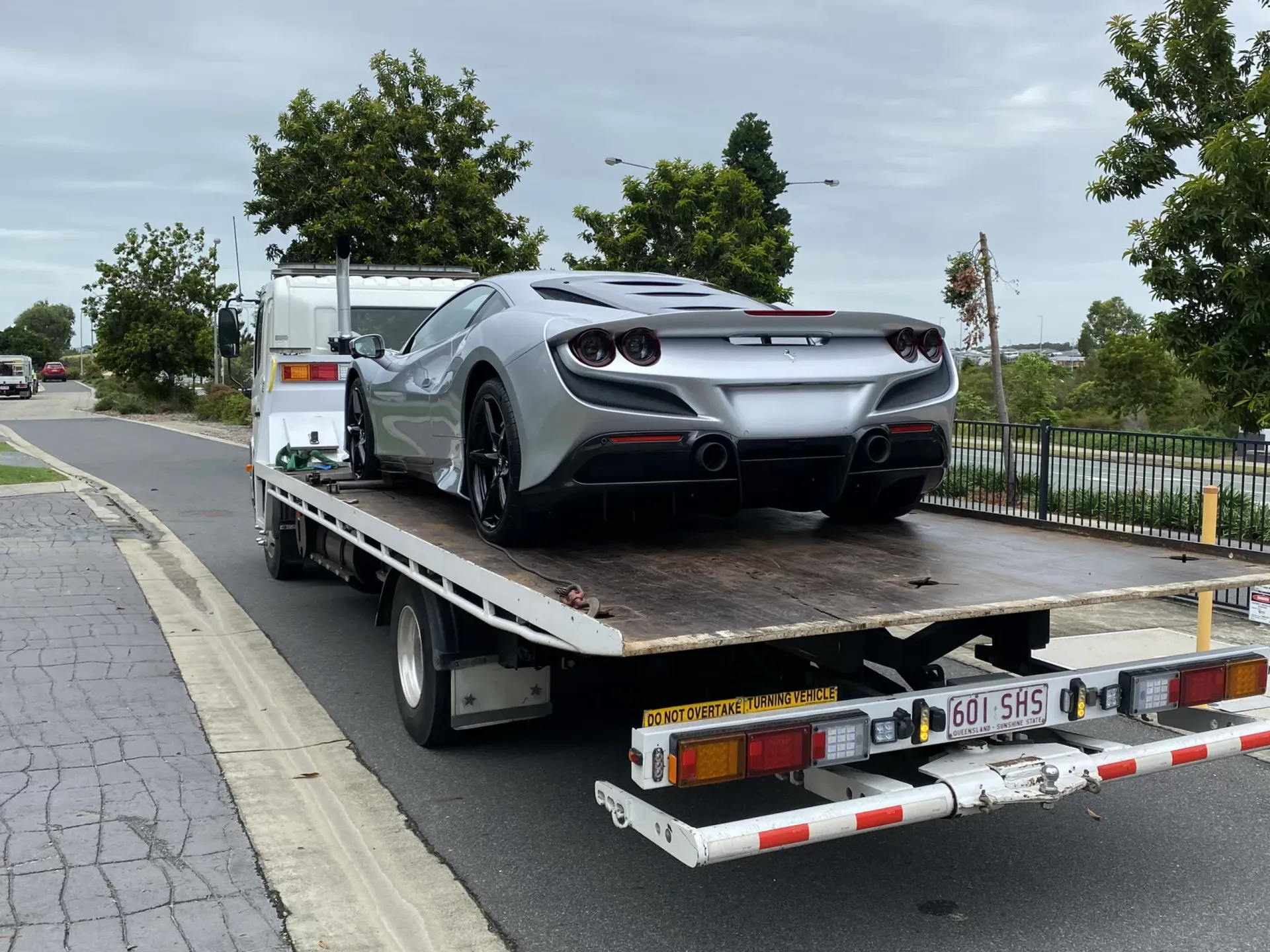 Silver sports car on a flatbed tow truck. Asphalt road with trees and a fence in the background.