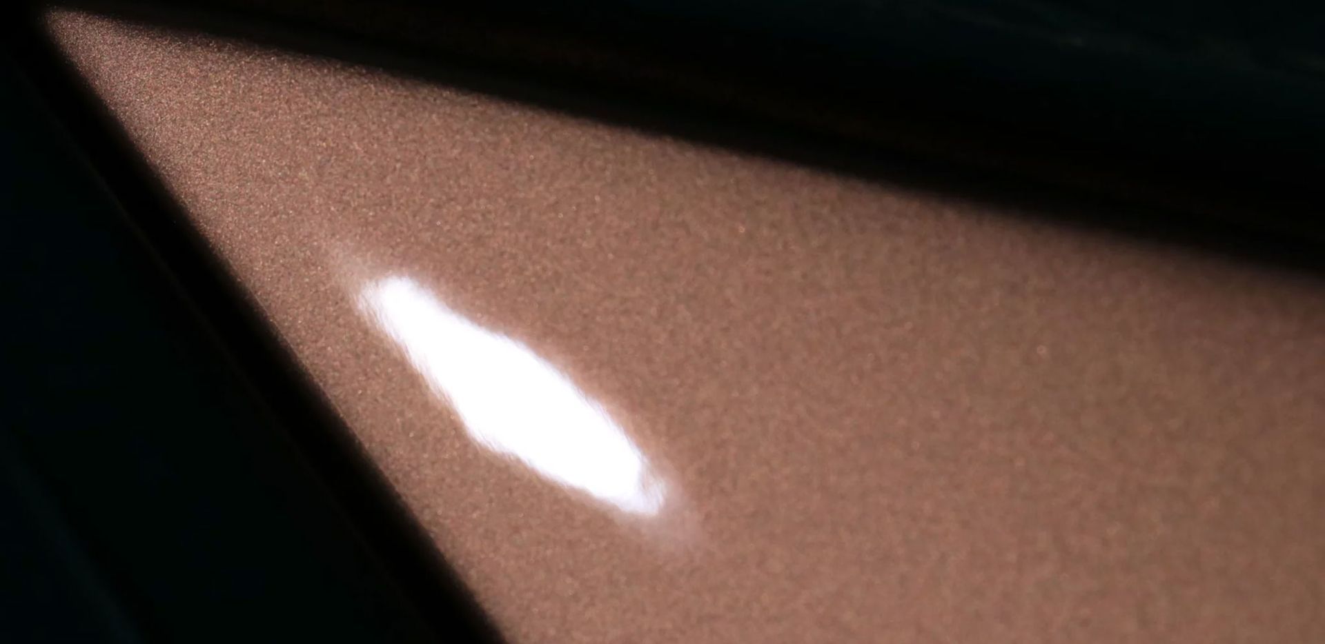 Close-up of a bronze-colored car panel with a bright, elongated reflection. Dark shadow in the corner.