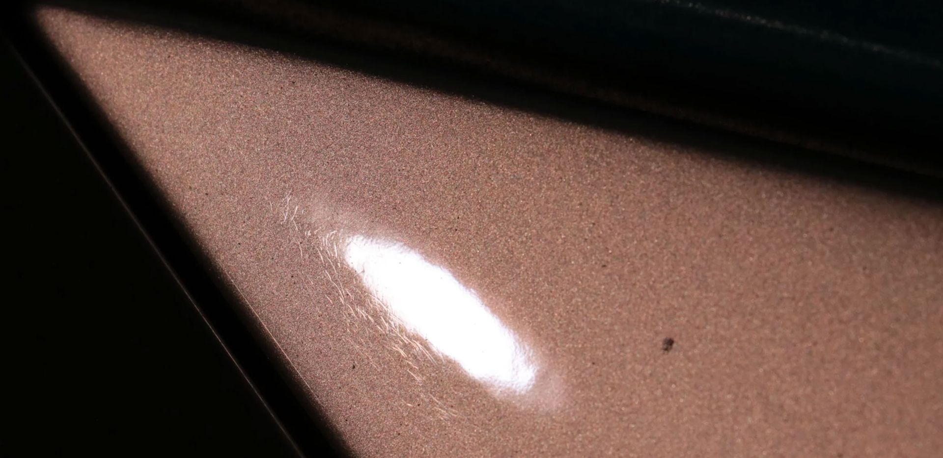 Close-up of a car's metallic brown paint surface with a bright white reflection and a small dark speck.