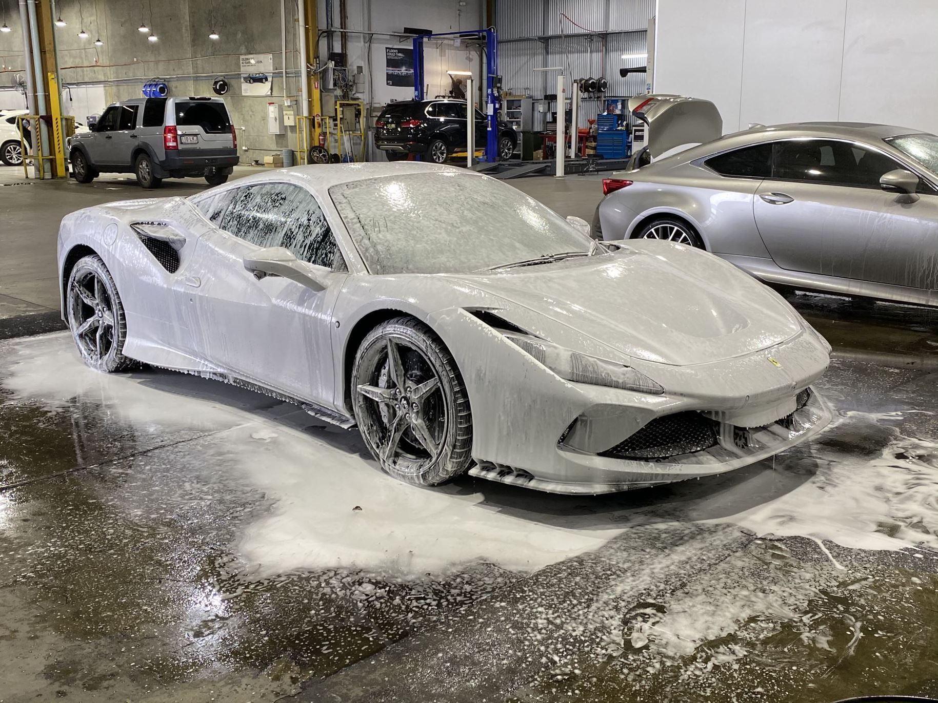 A white sports car covered in soap suds at a car wash. Another car is partially visible on the right.