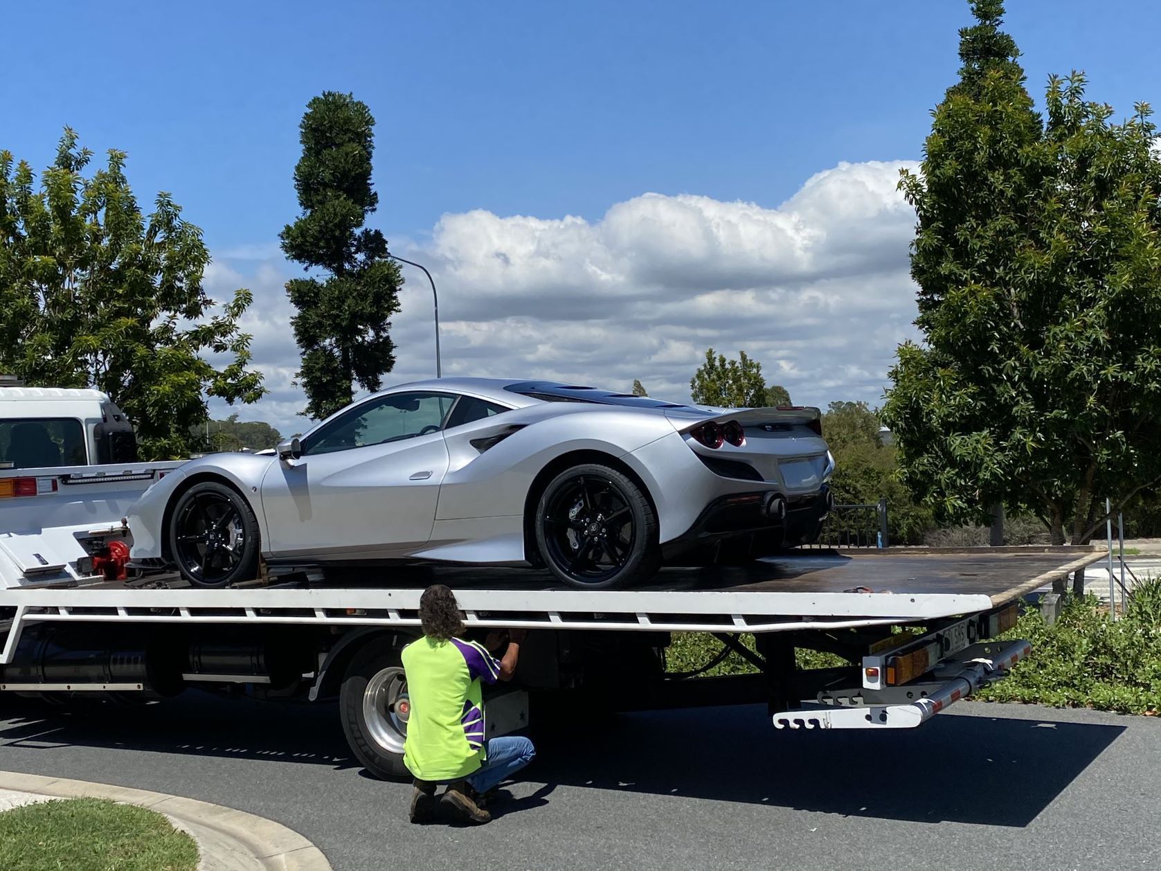 Silver sports car being loaded onto a flatbed tow truck by a person in a safety vest, outdoors on a sunny day.