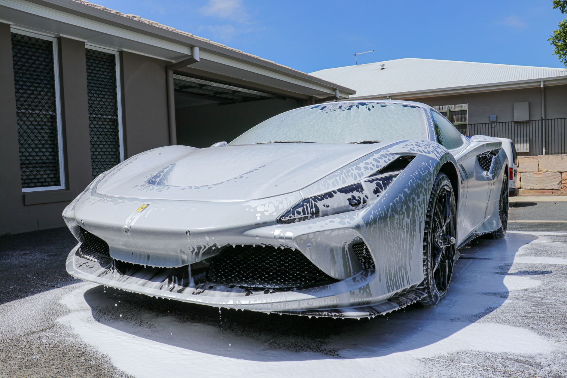 Silver Ferrari being washed with foamy soap in a driveway on a sunny day.