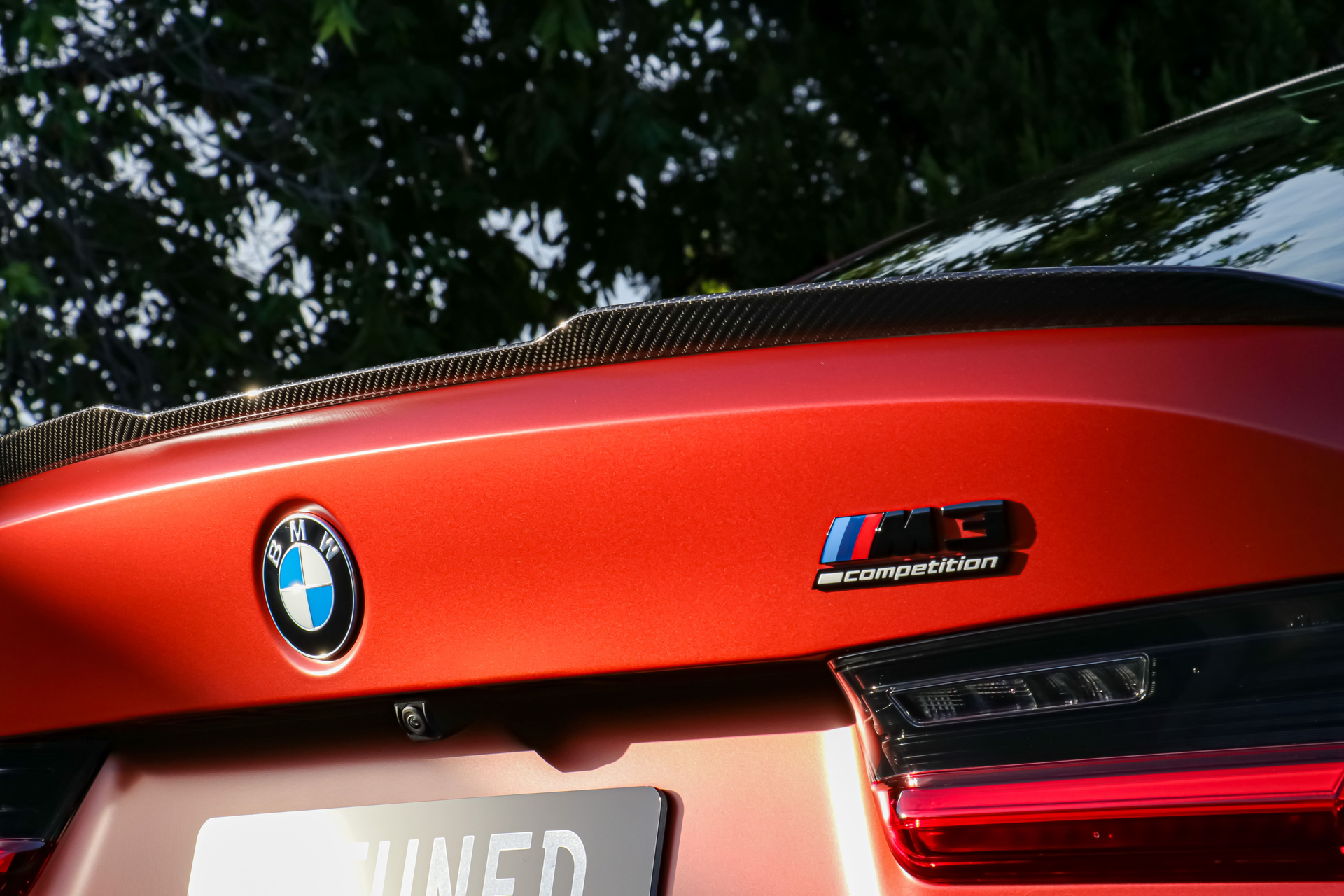 A close-up of a matte orange BMW M3 Competition's trunk, featuring the emblem, roundel logo, and carbon fiber spoiler.