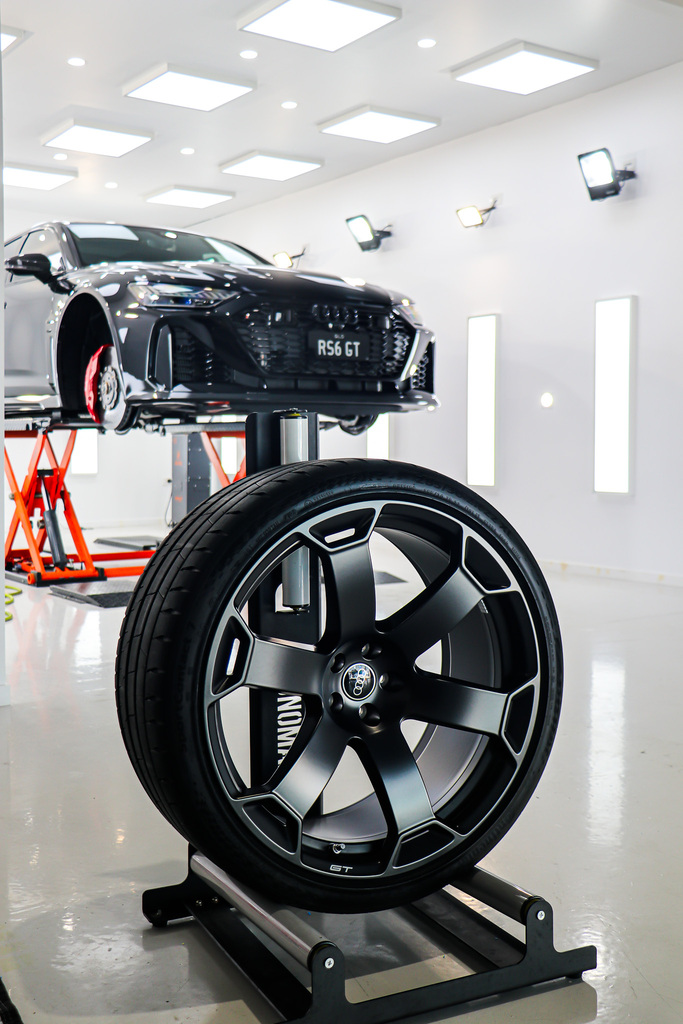 A black car wheel on a stand in the foreground, with a car raised on a lift in a bright, modern auto workshop.