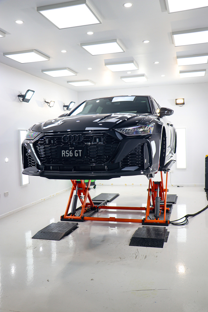 A black Audi RS6 GT car sits elevated on a bright orange hydraulic lift in a sterile, well-lit professional garage.