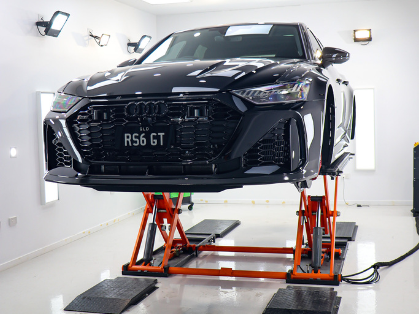 A black Audi RS6 GT car elevated on an orange scissor lift inside a bright, white-walled automotive workshop.