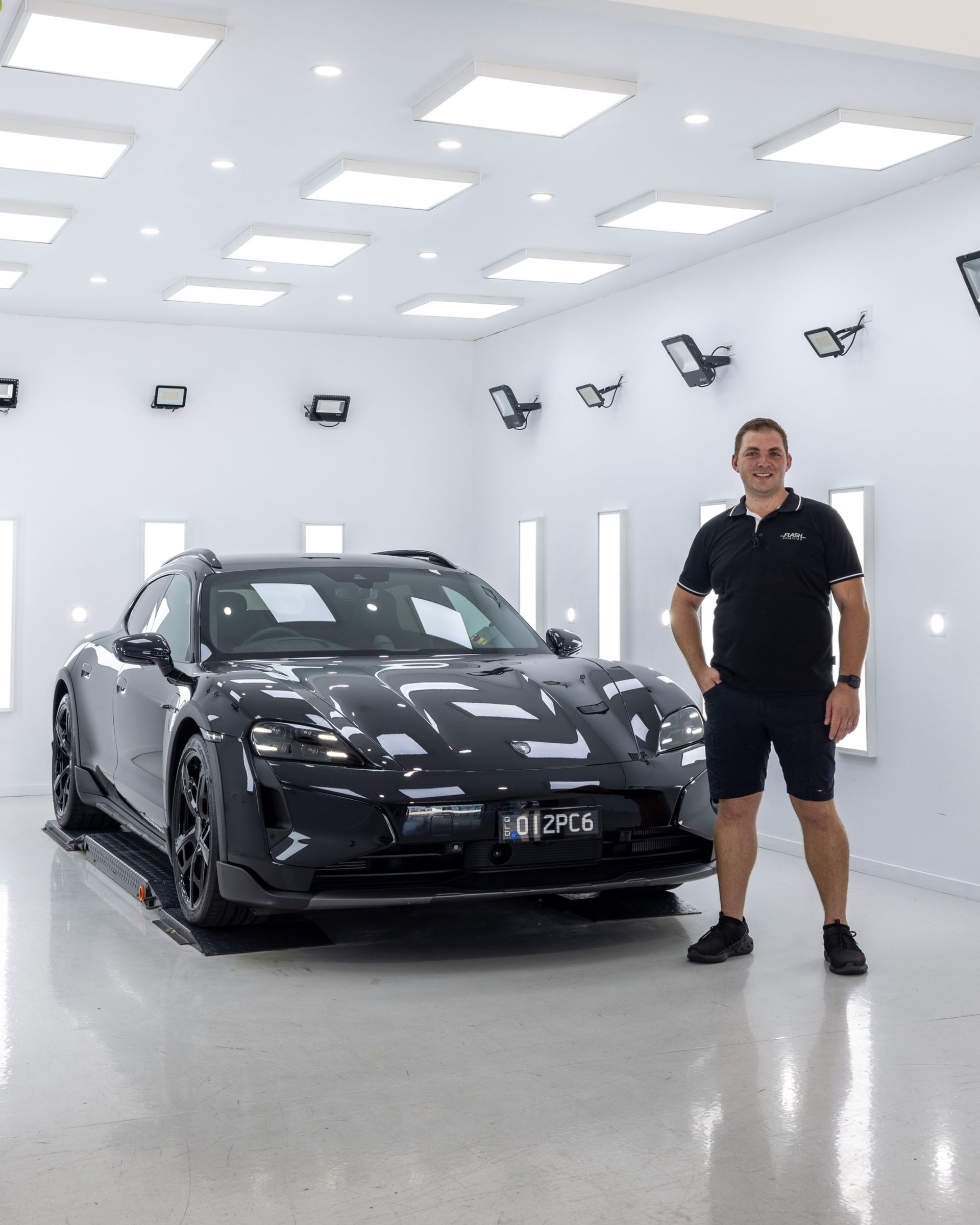 A person stands smiling next to a sleek black Porsche Taycan Cross Turismo in a brightly lit, white automotive studio.