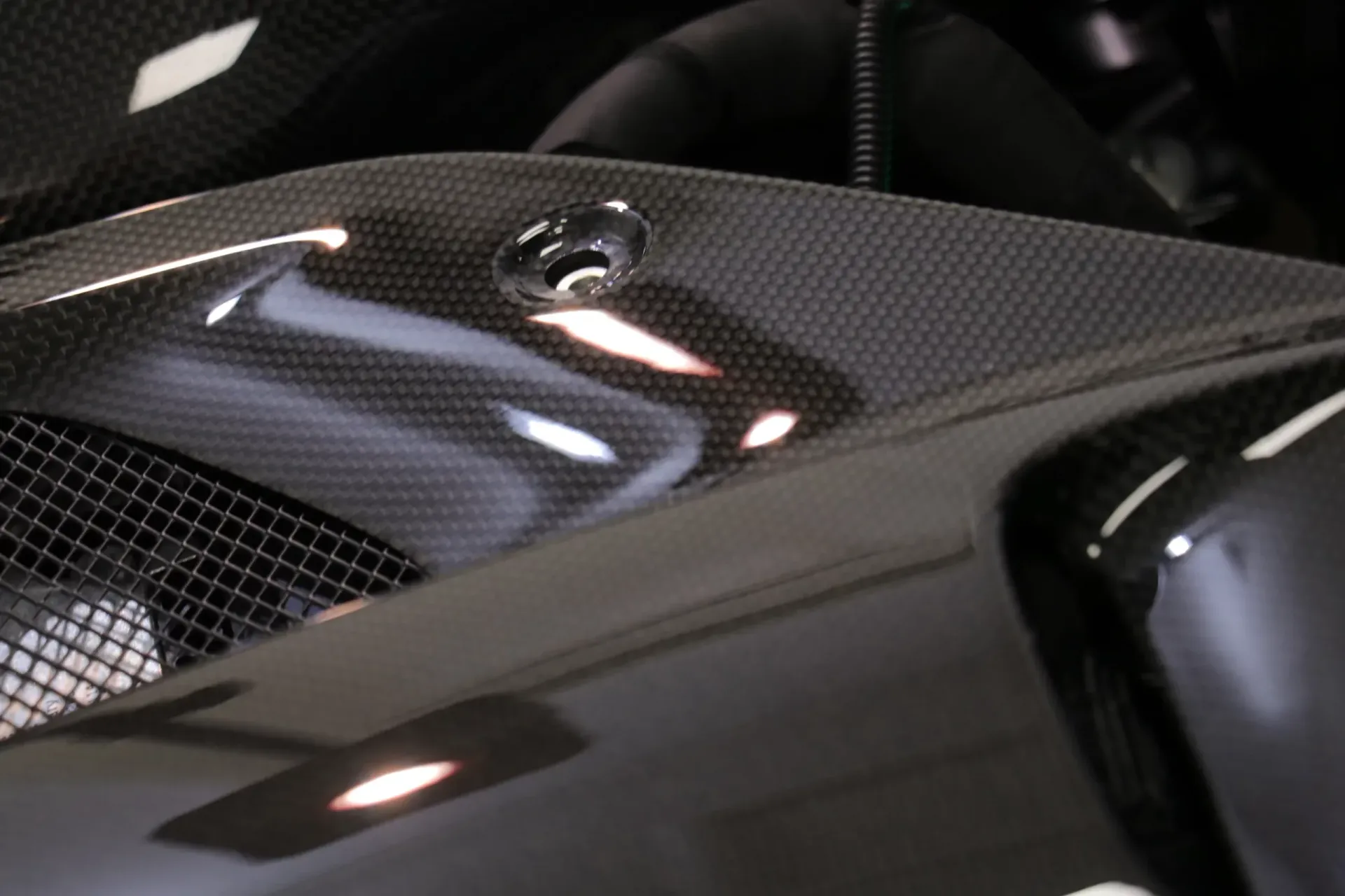 Close-up of a glossy, dark carbon fiber vehicle component. A silver fitting is set in the carbon fiber weave.