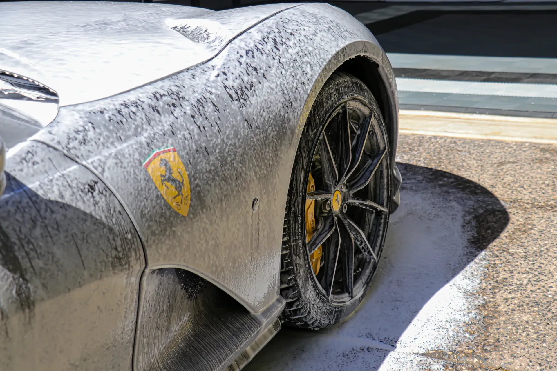A silver Ferrari being washed, covered in white foam; yellow Ferrari emblem visible.