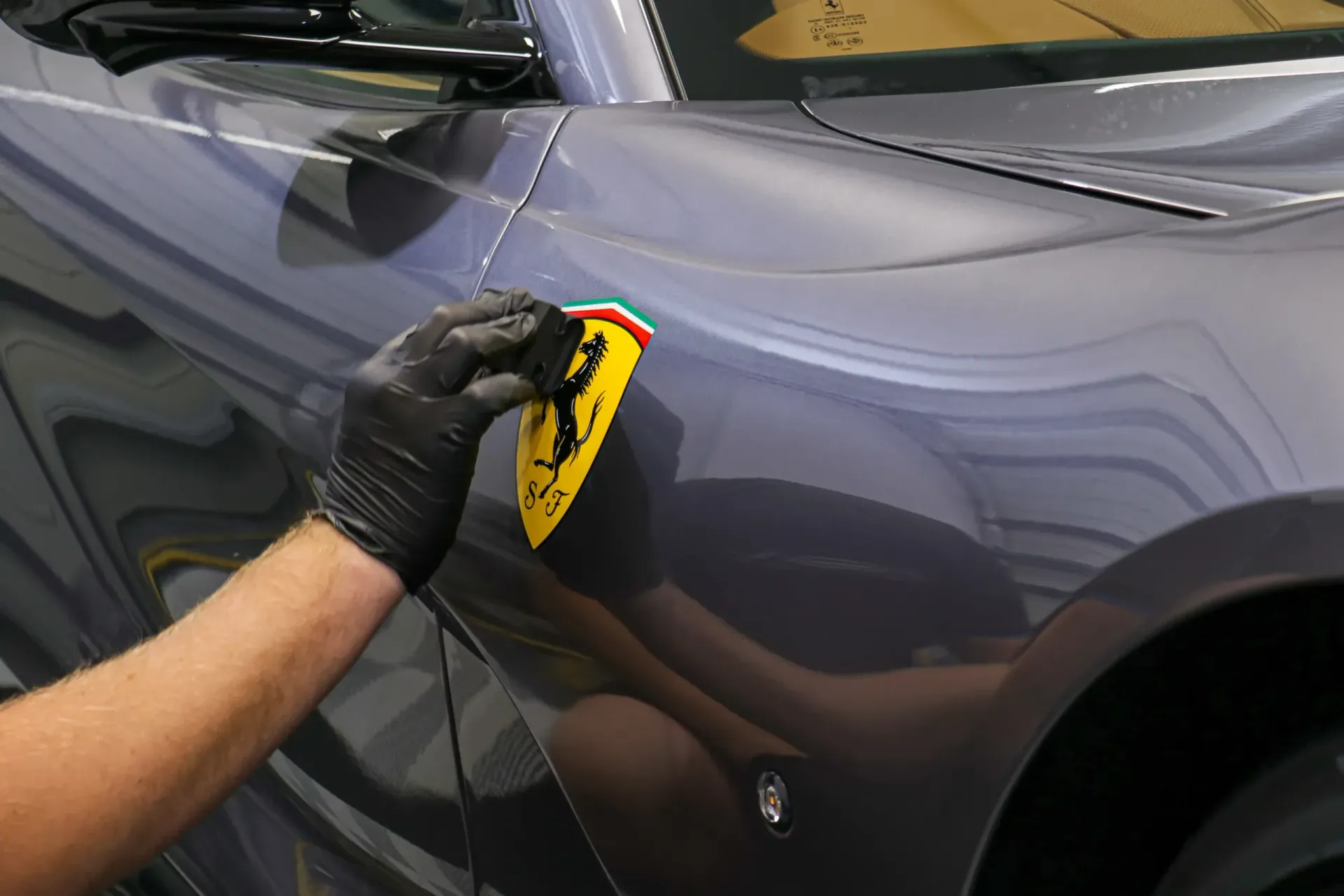 Person attaching a Ferrari emblem to a gray car's side panel, wearing a black glove.