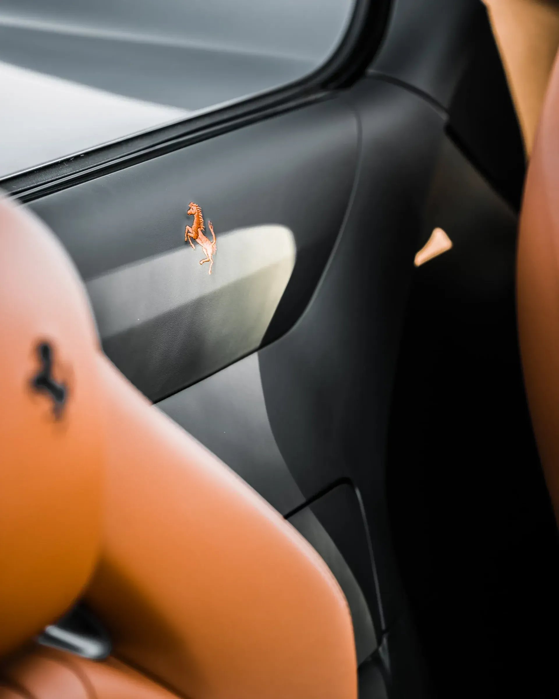 Interior detail of a Ferrari car with the iconic prancing horse logo on black leather. Brown seat is also visible.