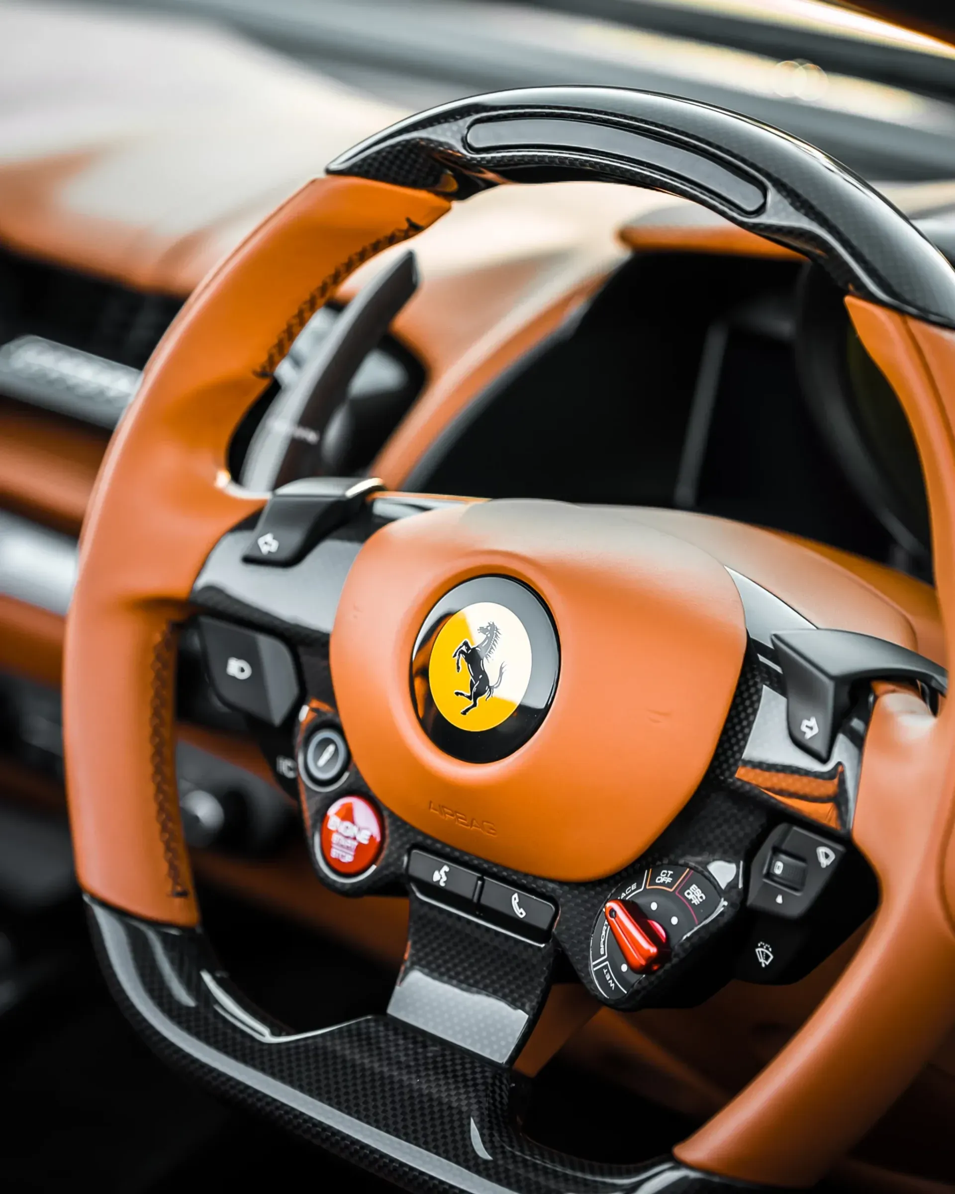 Close-up of a Ferrari steering wheel; orange leather with black carbon fiber accents and a red start button.