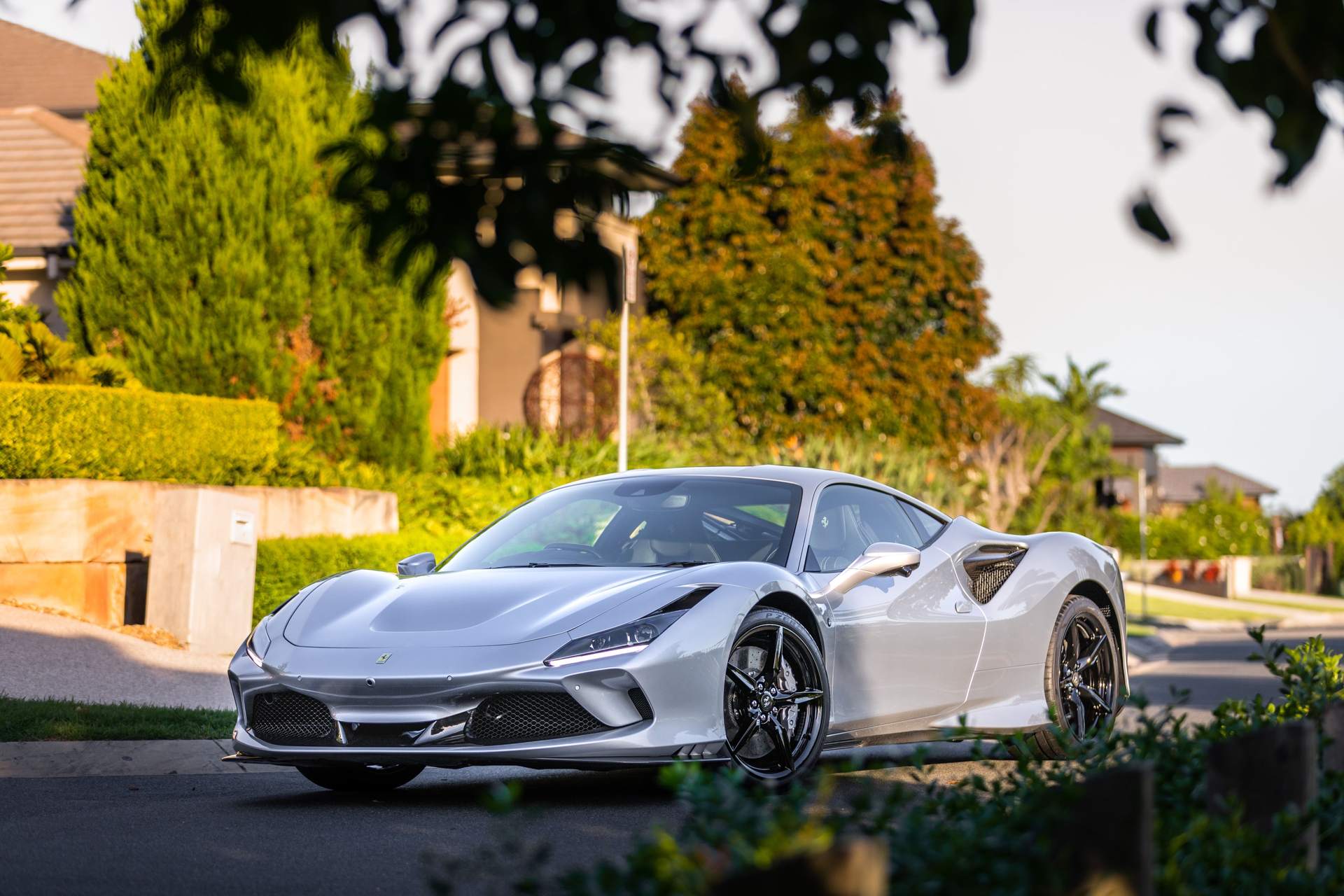 Silver Ferrari F8 Tributo sports car parked on a suburban street, trees and houses in the background.