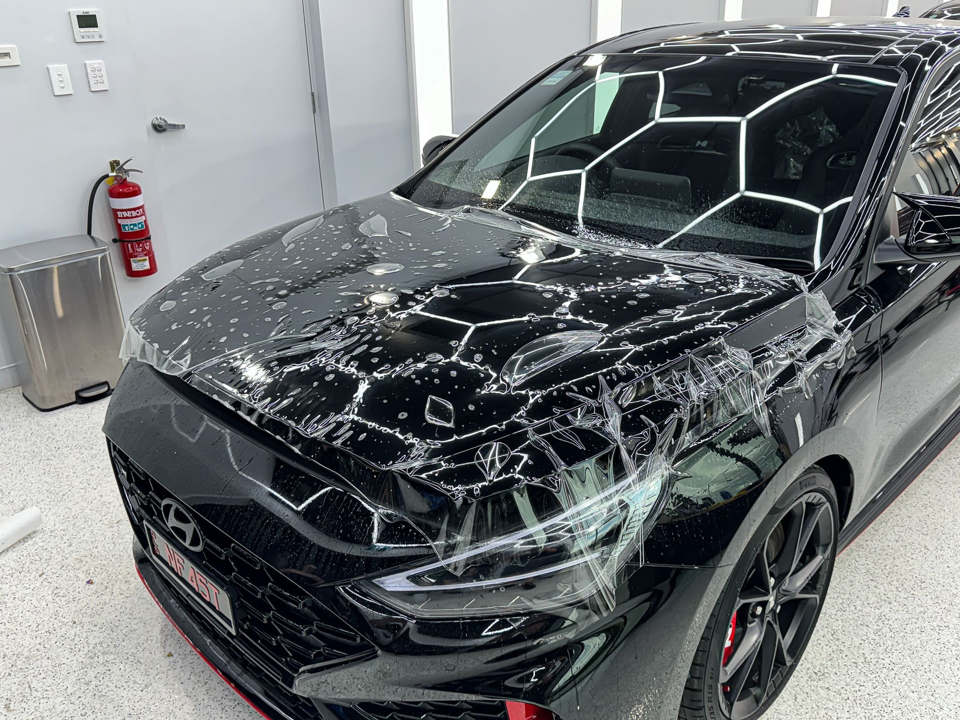 Black car with protective film applied to the hood in a well-lit shop.