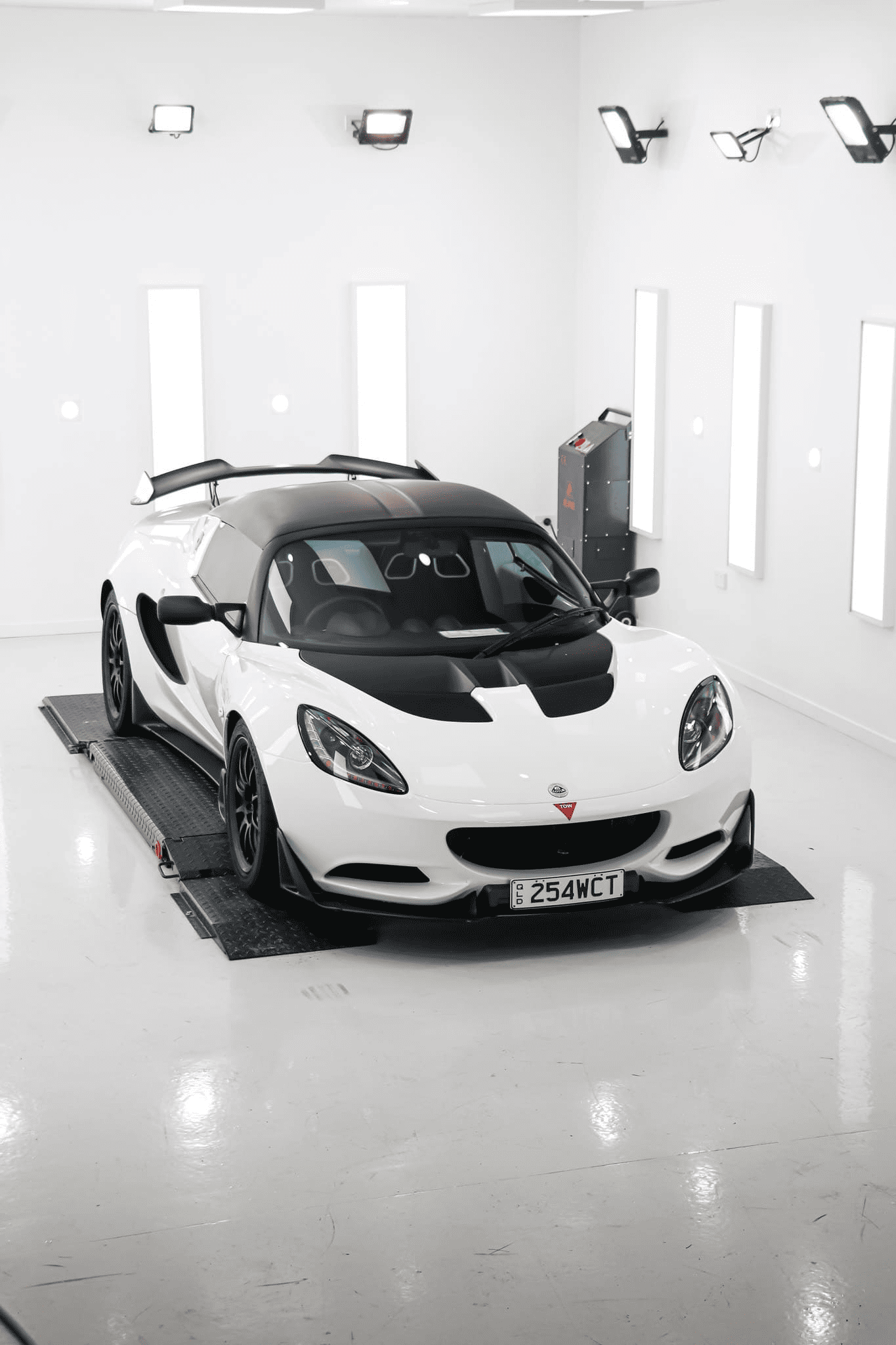 A white Lotus sports car with a black roof and hood parked inside a bright, white studio with overhead spotlights.