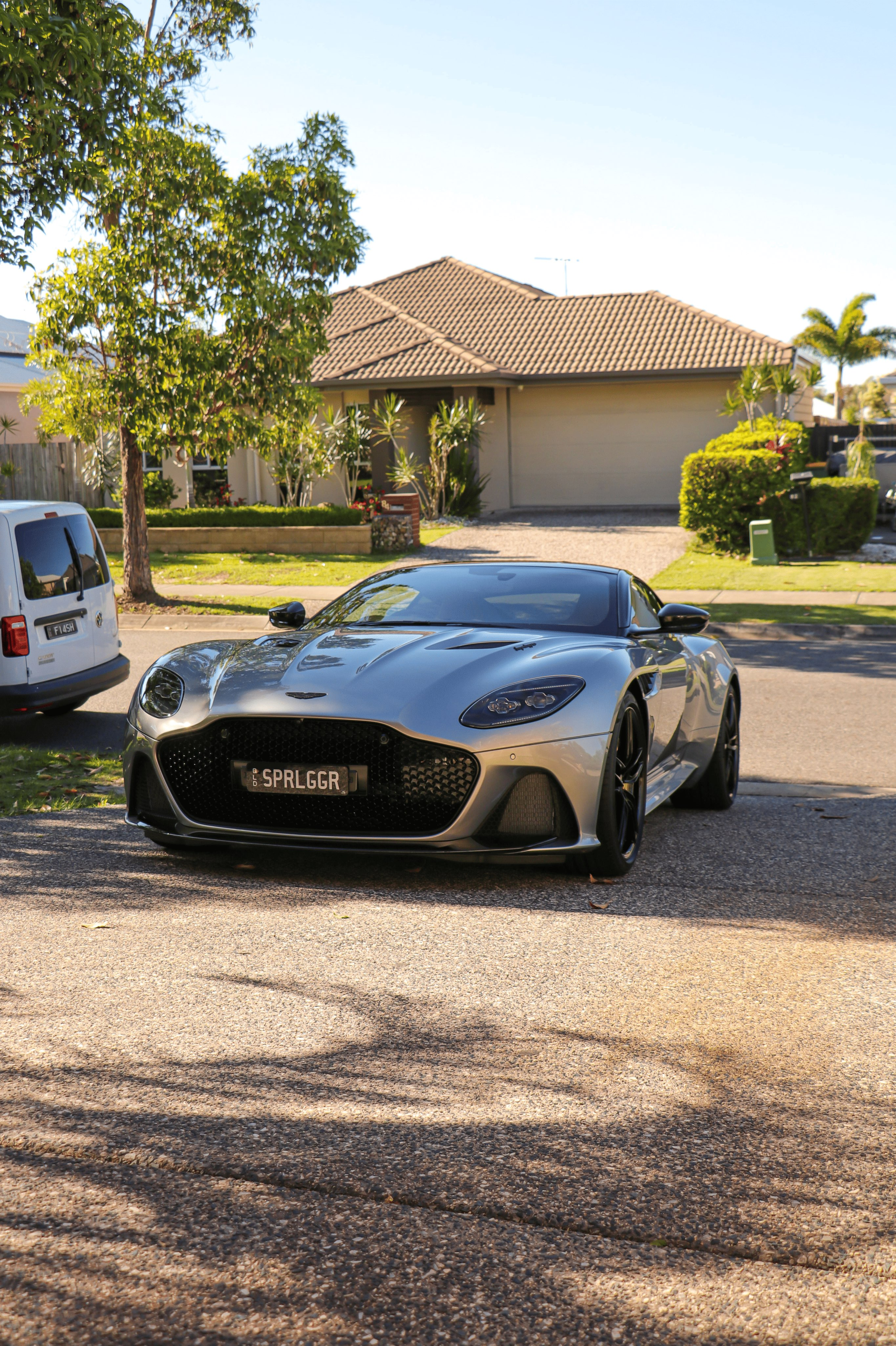 A silver Aston Martin sports car parked on a gravel driveway in front of a house on a sunny day.