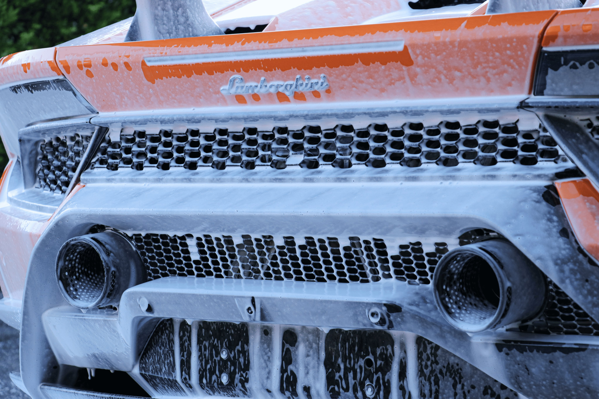 Close-up of the rear bumper of an orange Lamborghini covered in thick white car wash soap and foam.