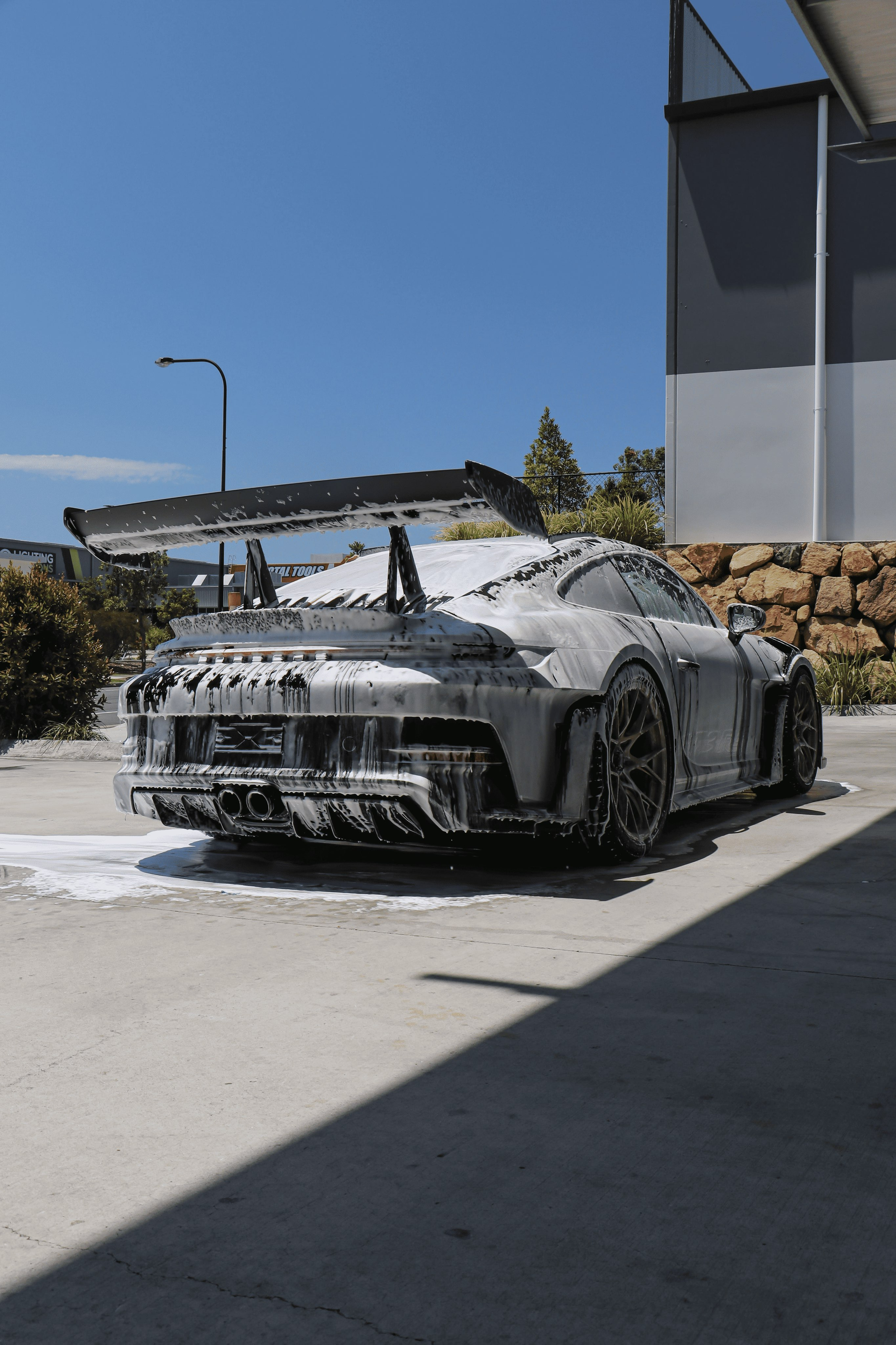 Porsche sports car covered in white foam being washed outdoors on a sunny day.