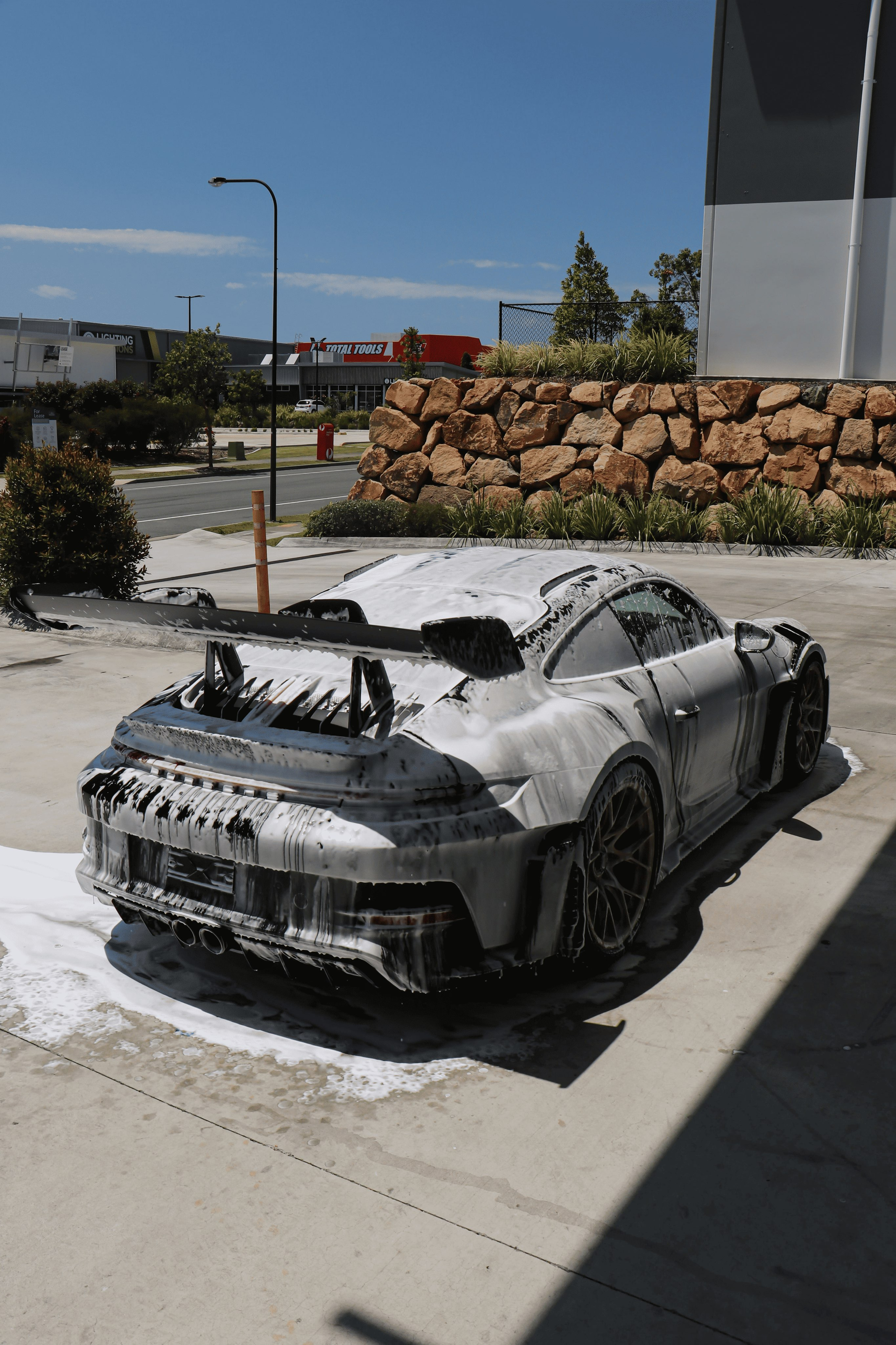 A silver sports car covered in white foam at a car wash on a sunny day.