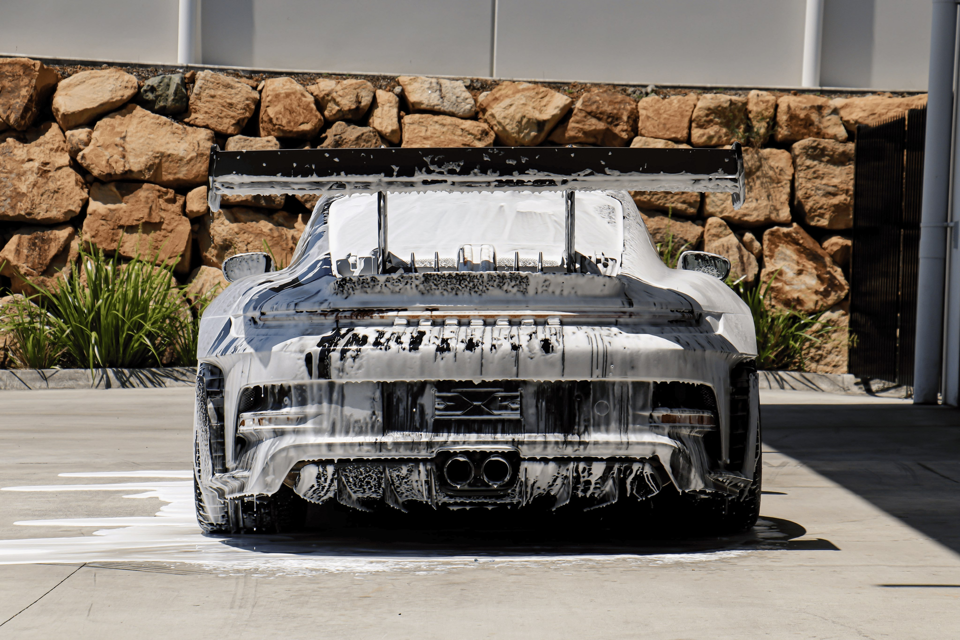 A white car covered in thick soap suds is parked on a concrete driveway in front of a stone wall.