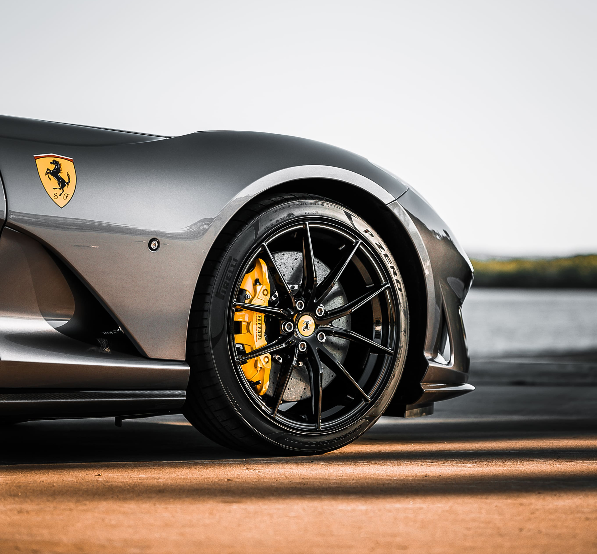 Gray Ferrari convertible parked on a road beside a body of water with trees in the background.